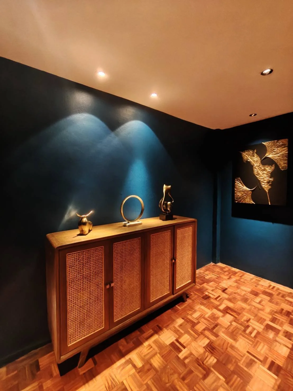 A wooden cabinet with woven doors stands against a dark teal wall, decorated with three art pieces, illuminated by ceiling lights, and a framed artwork showing gold leaves on a dark background.