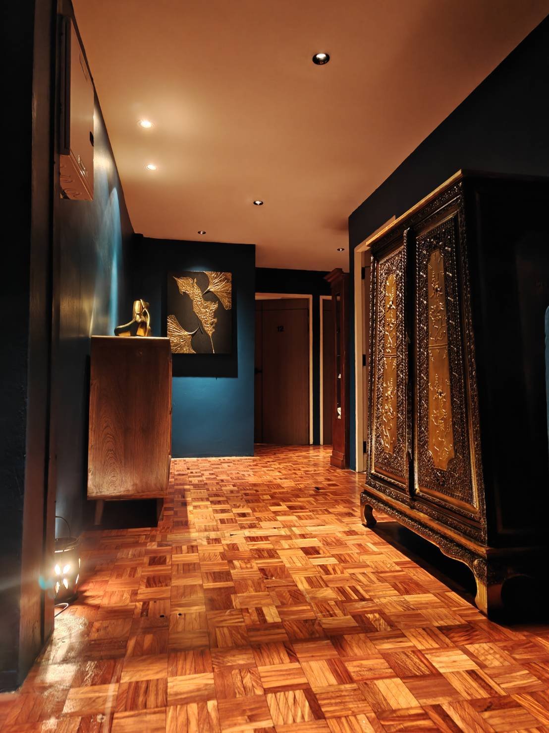 A hallway with hardwood flooring, dark blue walls, and ceiling lights. On the left, there is a wooden console table with a decorative sculpture, and on the right, a large ornate black and gold cabinet. The hallway leads to a closed door at the back.