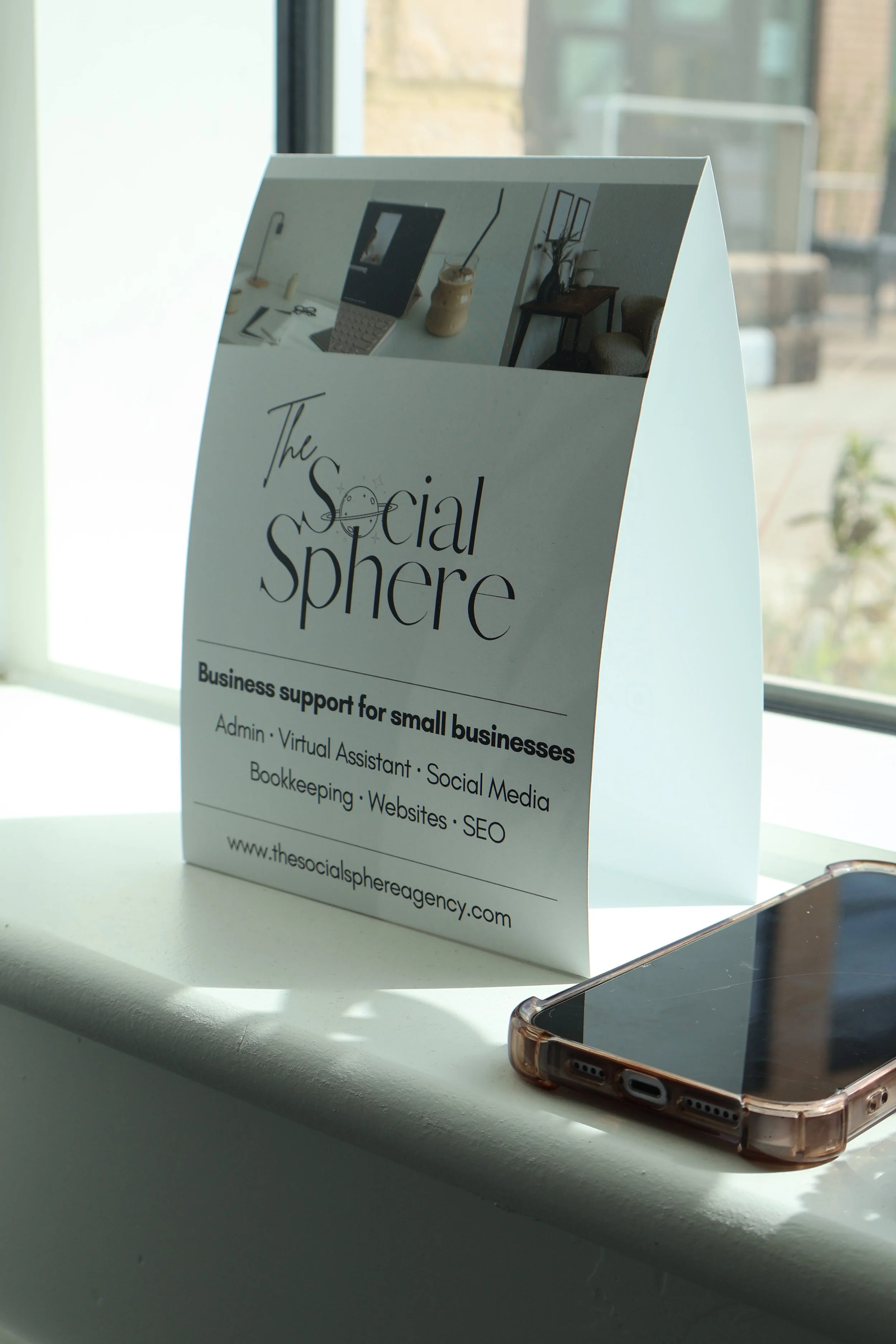 A white promotional sign on a windowsill advertising social media and business support services for small businesses. The sign shows images of an office workspace and contains text about the services offered, including admin, virtual assistant, social media, bookkeeping, websites, and SEO, with a website URL at the bottom.