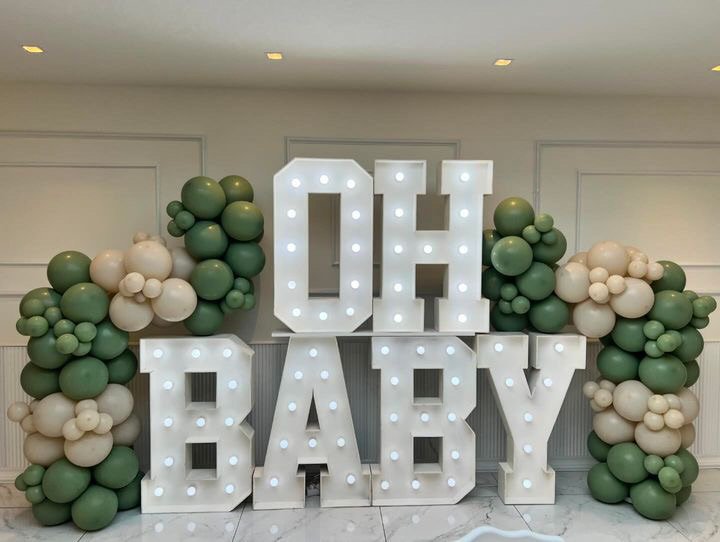 Verlichte letters met "OH BABY" omgeven door groene en beige ballonnen.