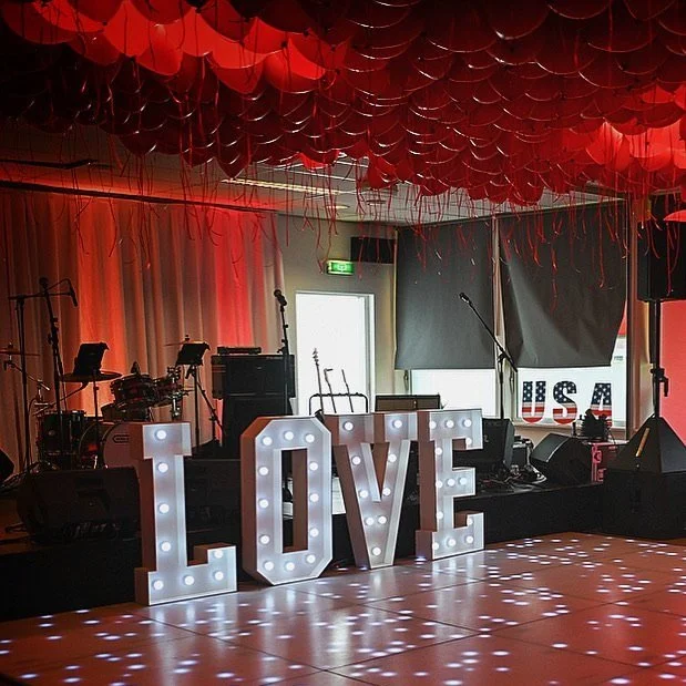 Decoratieve opstelling met grote verlichte letters 'LOVE' en muziekinstrumenten op een podium. Plafond versierd met rode ballonnen en borden met de tekst 'USA'. De sfeer is feestelijk en romantisch.