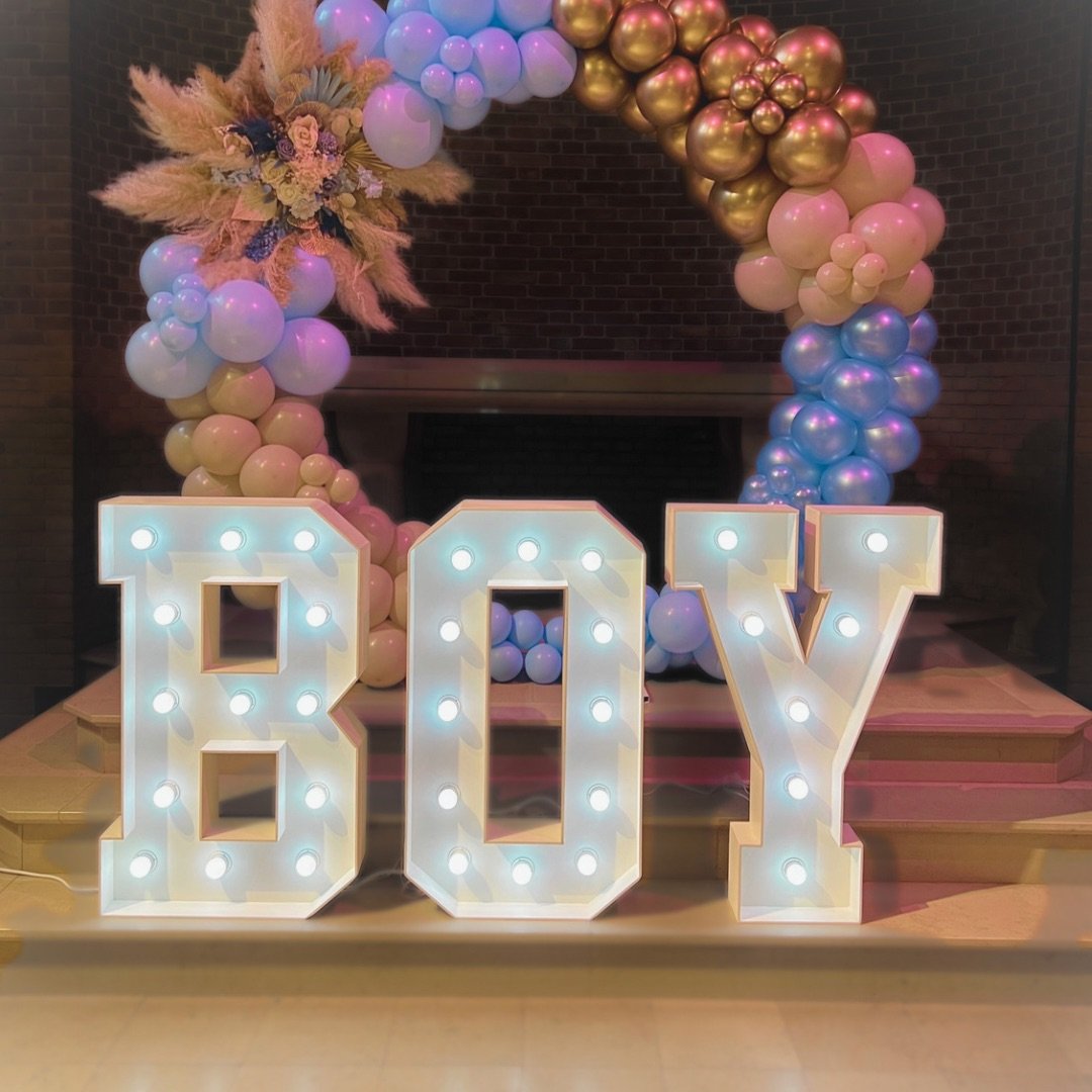 Lichtgevende letters "BOY" met een gebogen ballondecoratie in blauw, roze en goud, versierd met bloemen en pampagras.