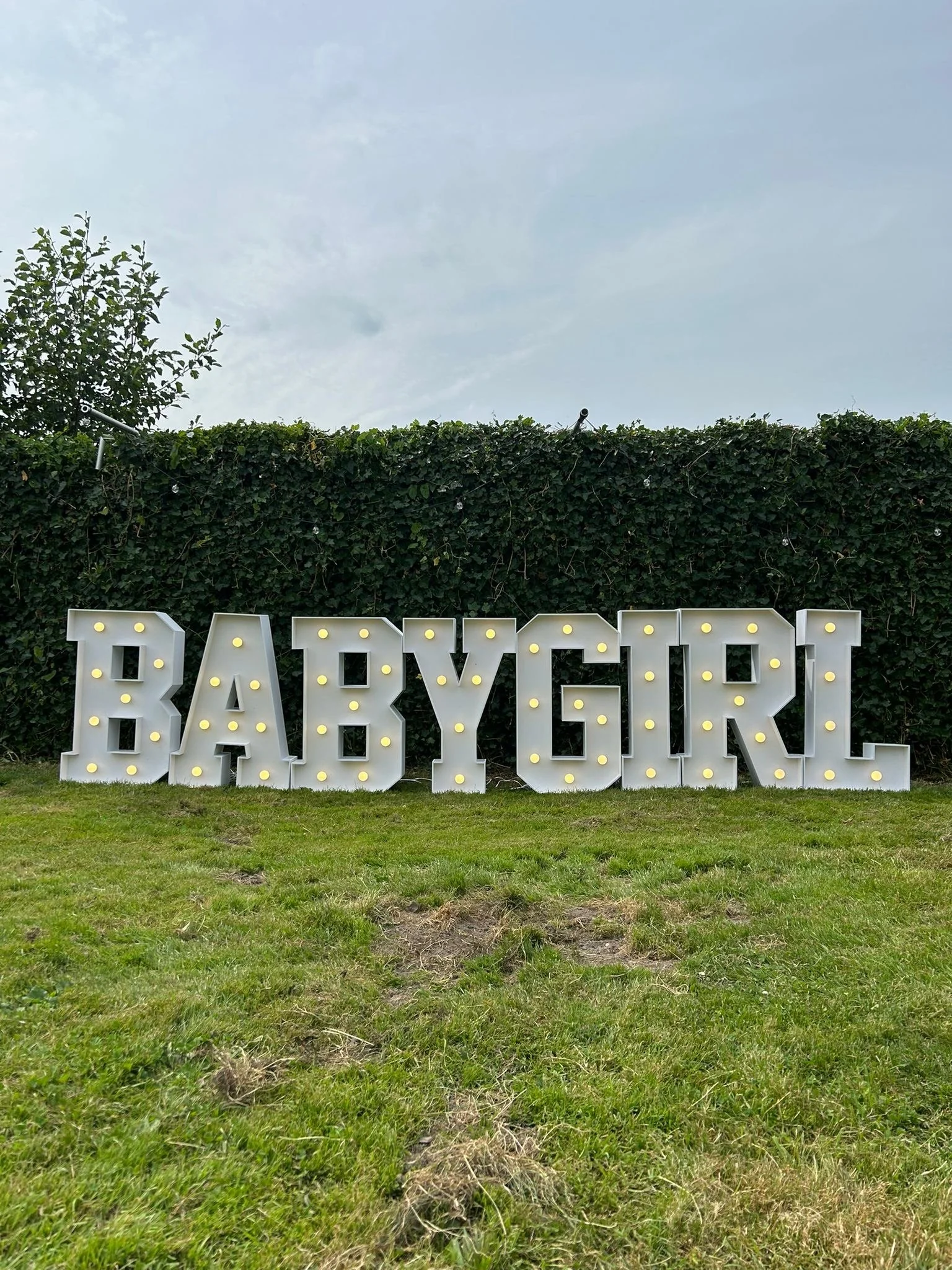 Grote verlichte letters in een tuin die 'BABYGIRL' spellen voor een heg op een grasveld.