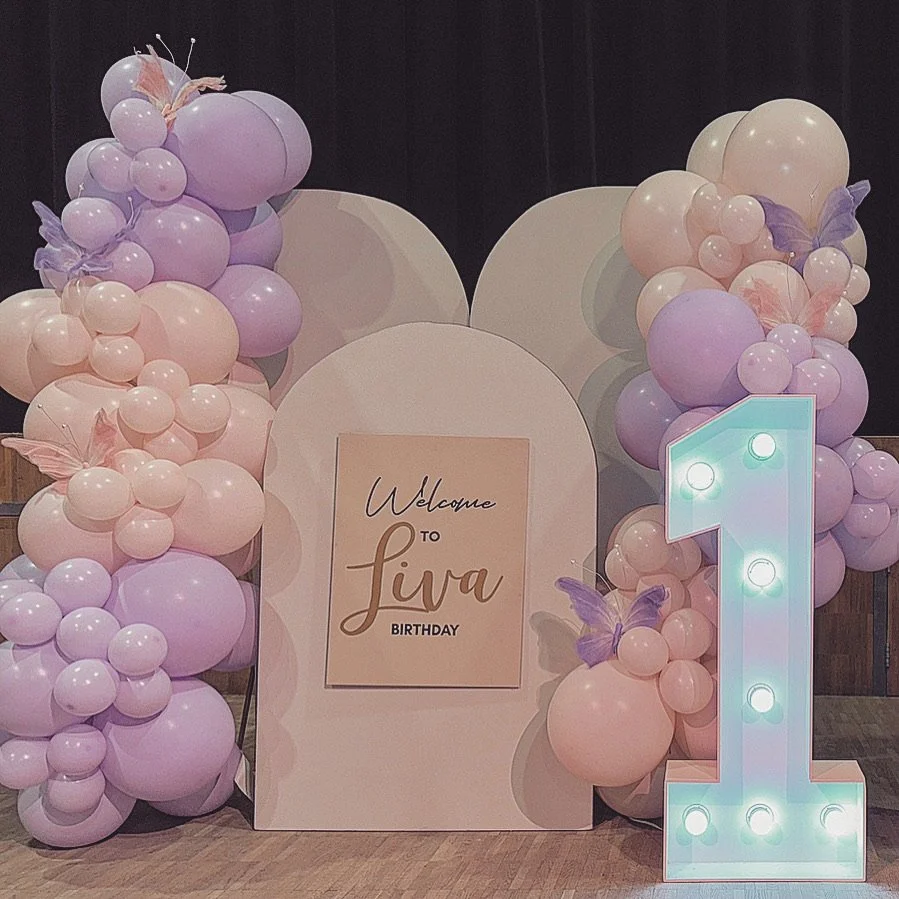 Verjaardagsdecoratie met ballonnen in pastelkleuren, een verlichte nummer 1, en een bord met de tekst "Welcome to Liva Birthday."