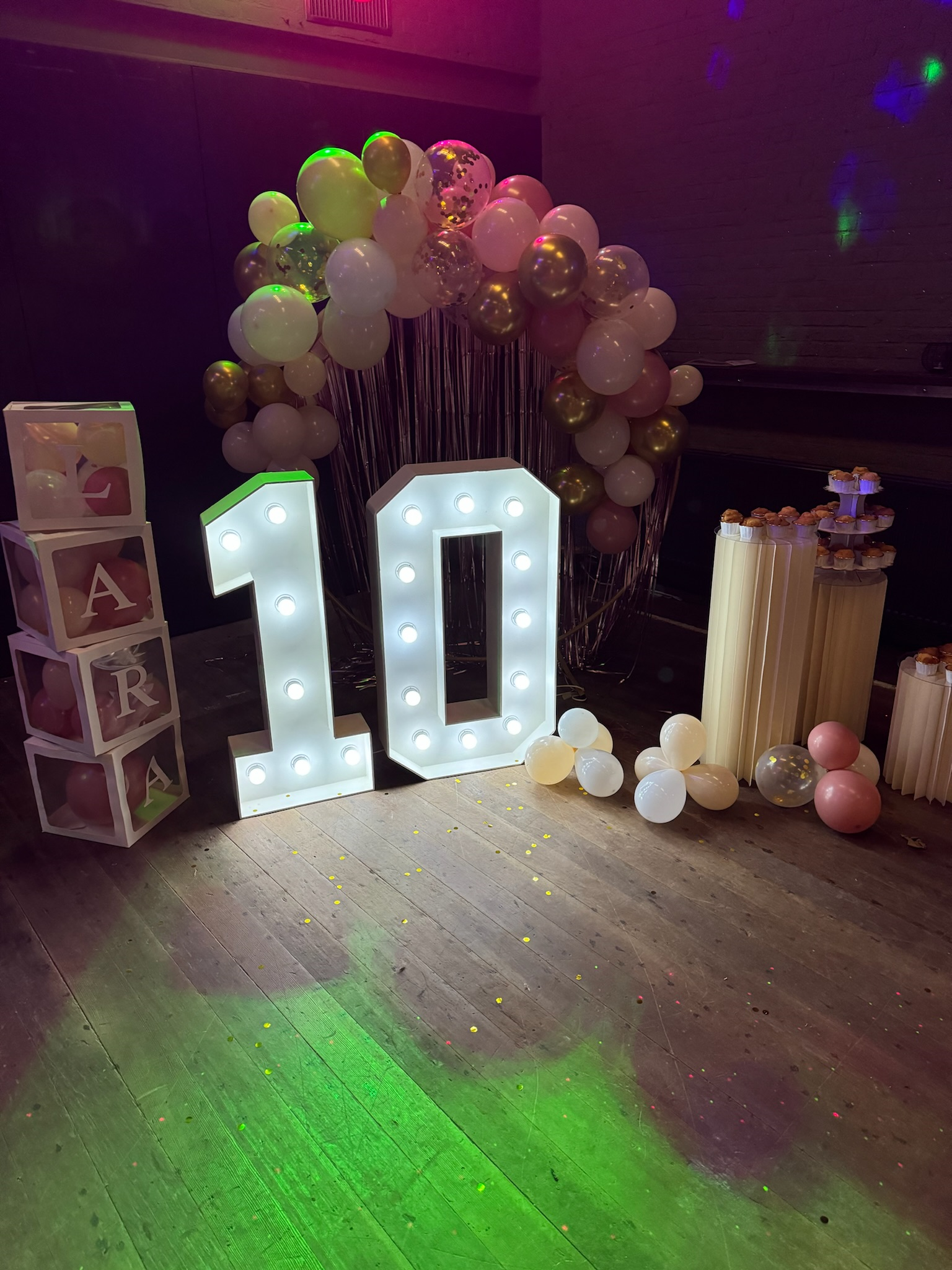 Feestdecoratie met een grote verlichte '10' en ballonnenboog op de achtergrond, met naastliggende decoratieve blokken met letters. Cupcakes staan op een tafel rechts.