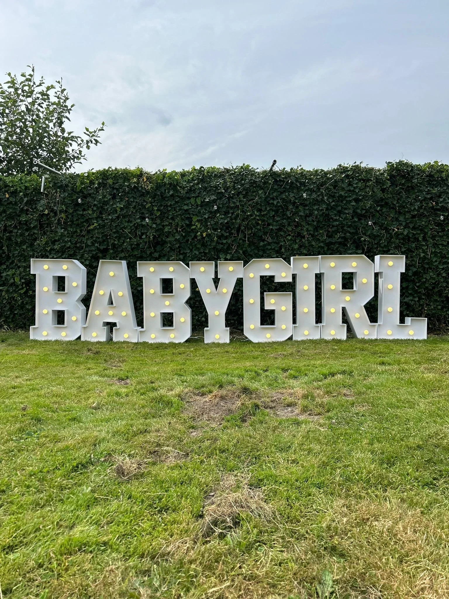 Grote verlichte letters die "BABYGIRL" spellen, geplaatst op een grasveld voor een heg.