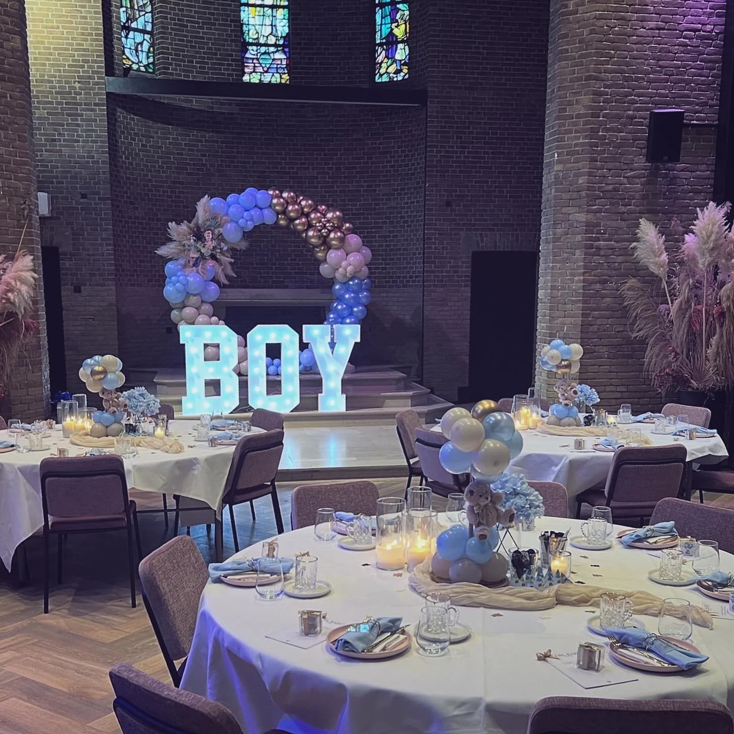 Decoratie voor een babyshower in een zaal met tafels, versierd met blauwe ballonnen en het woord 'BOY' in verlichte letters.