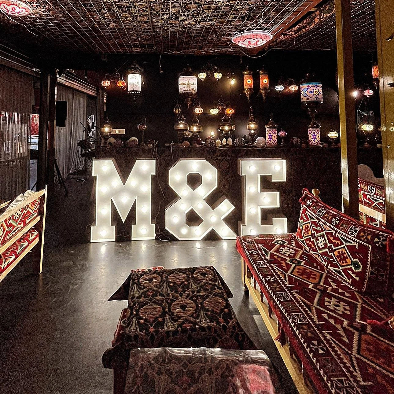 Feestzaal gedecoreerd voor een baby shower of gender reveal party, met ronde tafels gedekt met blauwe en gouden ballonnen, kaarsen en tafeldecoraties. In het midden is er een grote verlichte "BOY"-display met een boog van ballonnen in verschillende kleuren. De sfeer is feestelijk met gedempt licht en gekleurde verlichting.