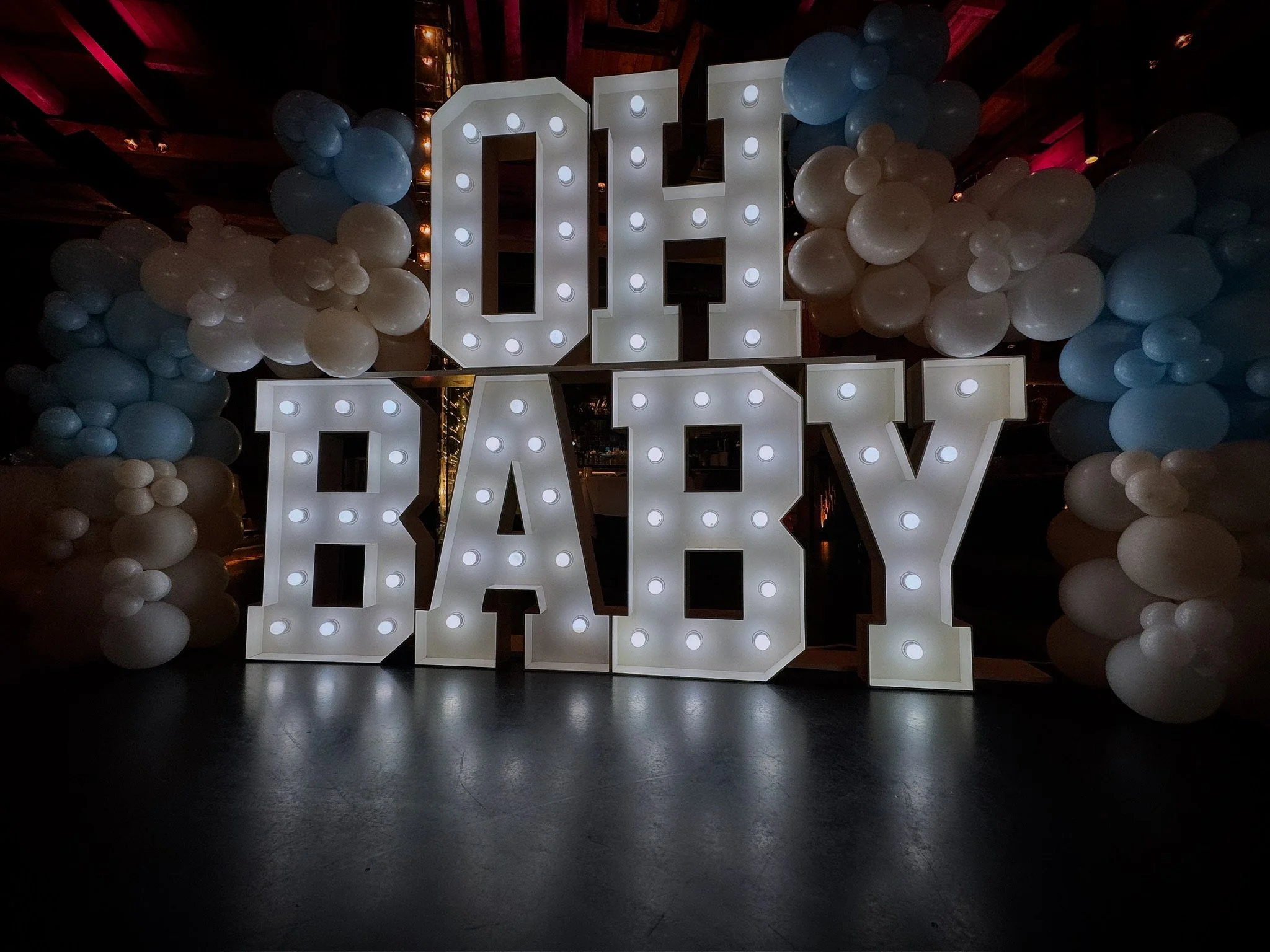 Verlichte letters "OH BABY" omringd door witte en blauwe ballonnen.