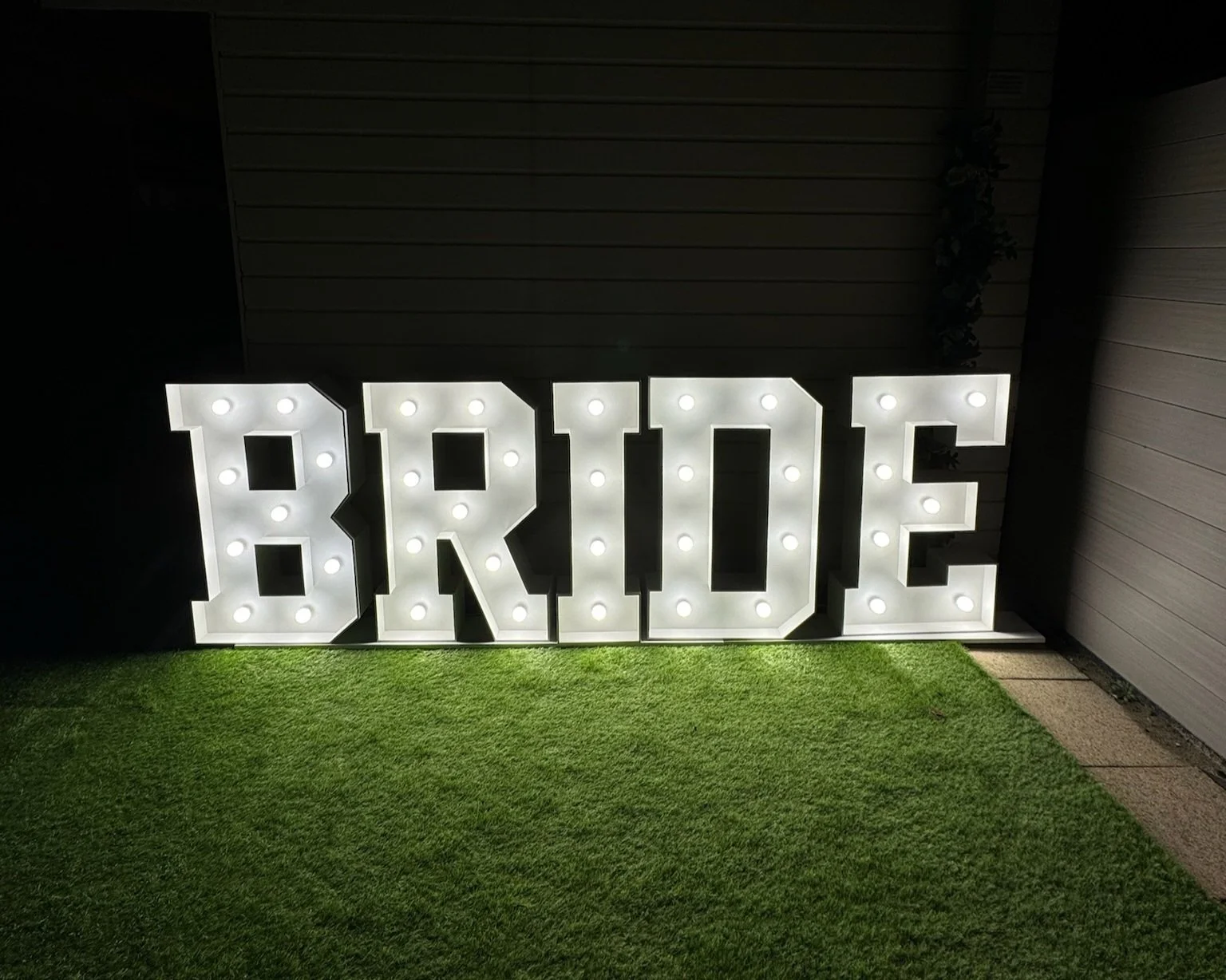 Verlichte letters vormen het woord 'BRIDE' op gras in de avond.