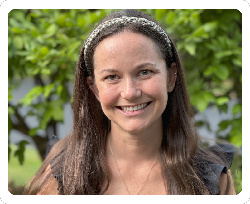 A woman smiling outdoors with green foliage in the background, wearing a headband and a black top.