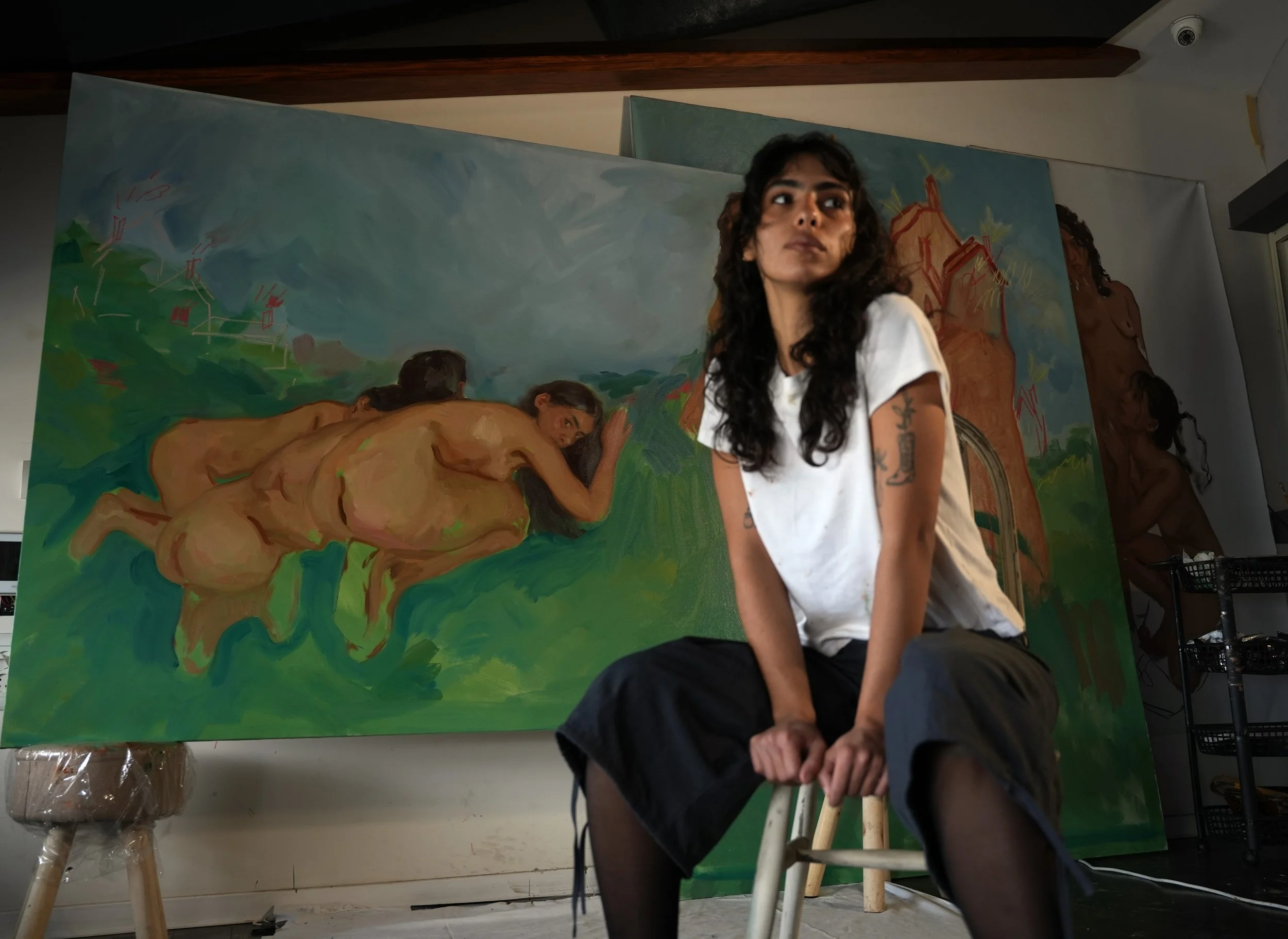 An artist with tattoos on her arm sitting on a stool in an art studio, with large colorful paintings of nude women in a landscape background behind her.