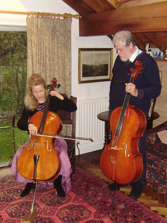 A woman and a man playing cellos indoors, near a window and a painting, with a red patterned rug on the floor.