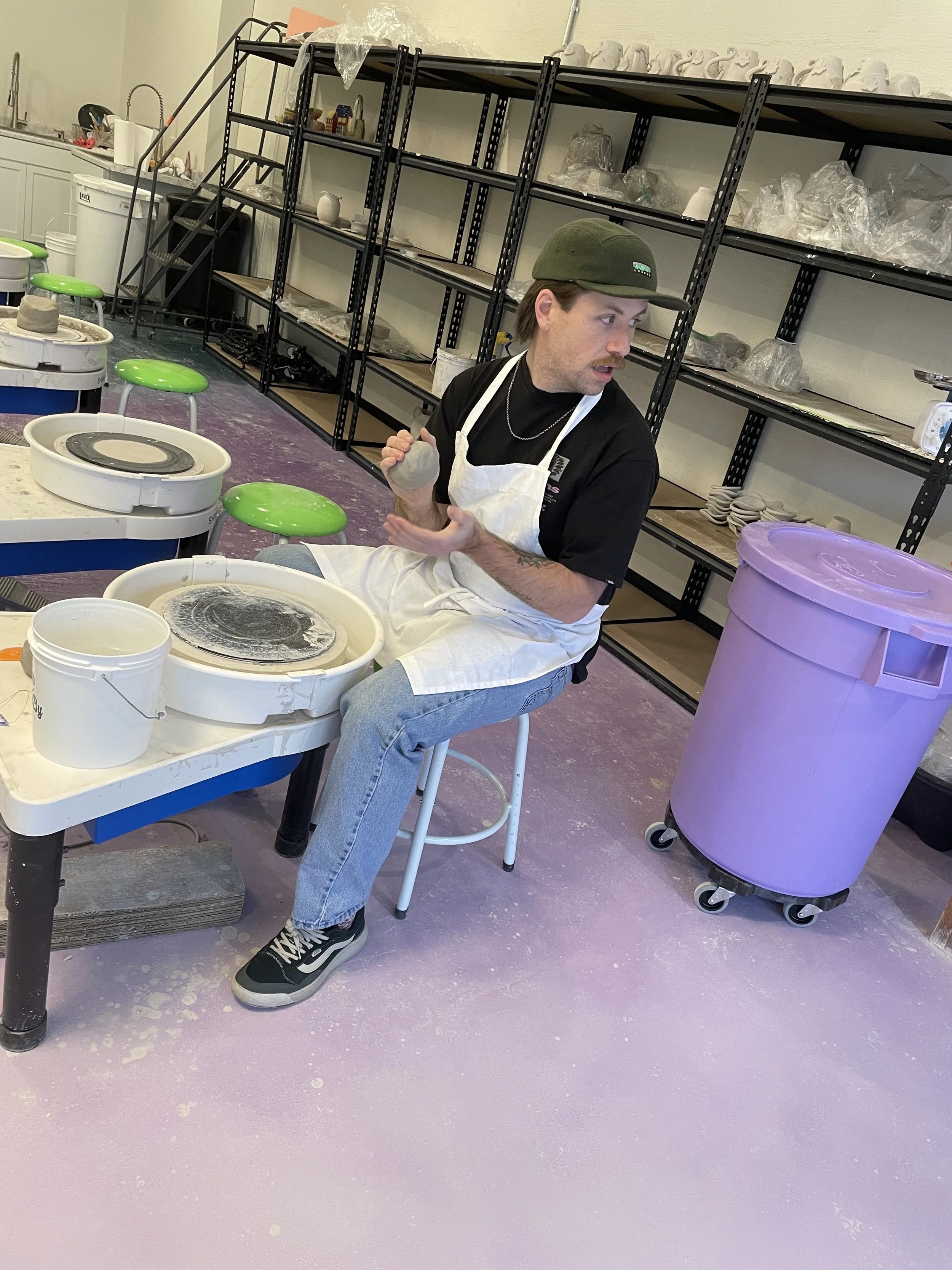 A man sitting on a stool in a ceramics studio, surrounded by pottery wheels, shelves with pottery supplies, and a large purple trash bin.