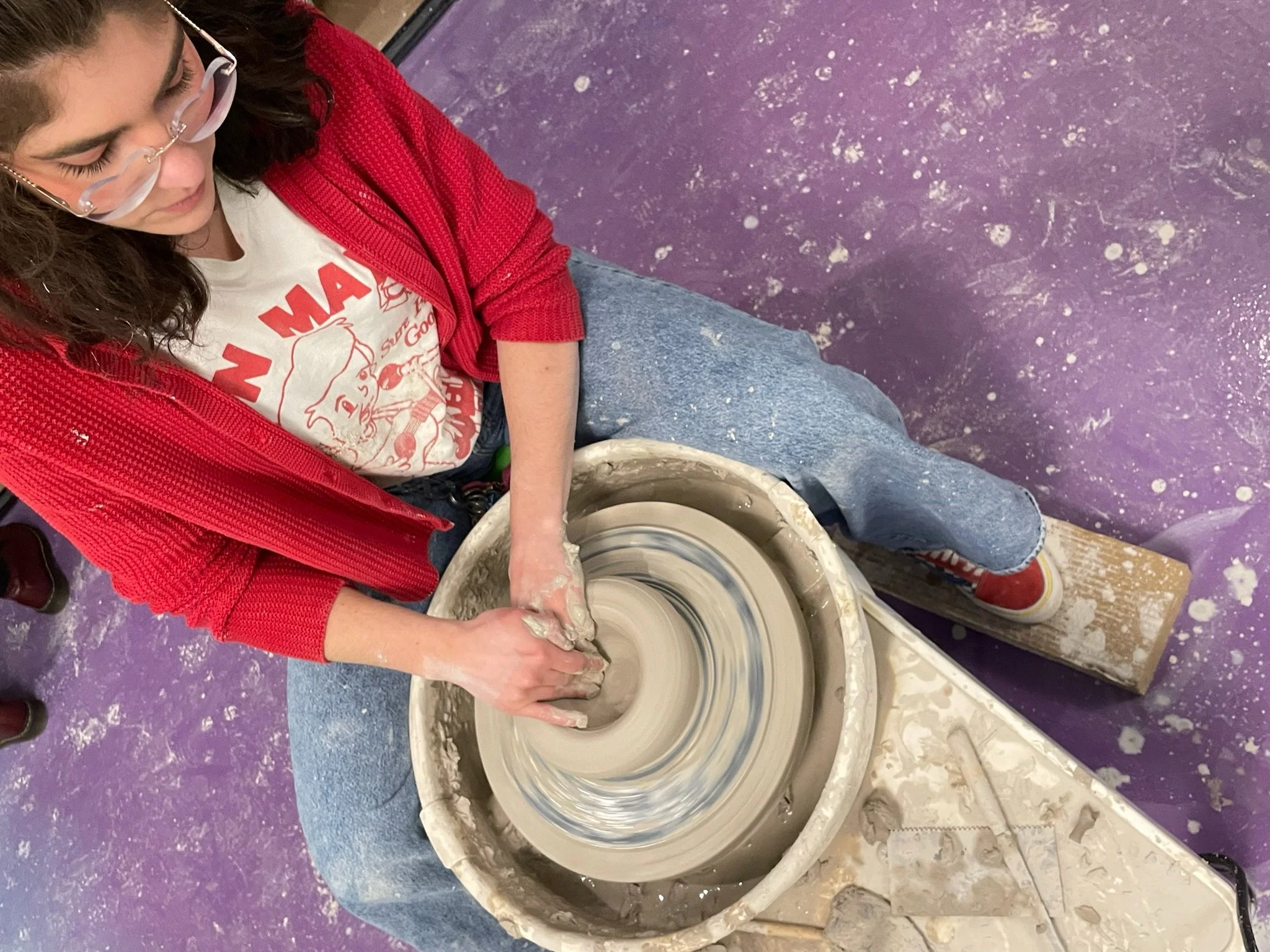 A young woman wearing glasses, a white T-shirt, a red jacket, and blue jeans is sitting at a pottery wheel, shaping wet clay with her hands. The pottery wheel spins as she works on her clay creation. The floor is purple and there is some pottery clay splattered on it.