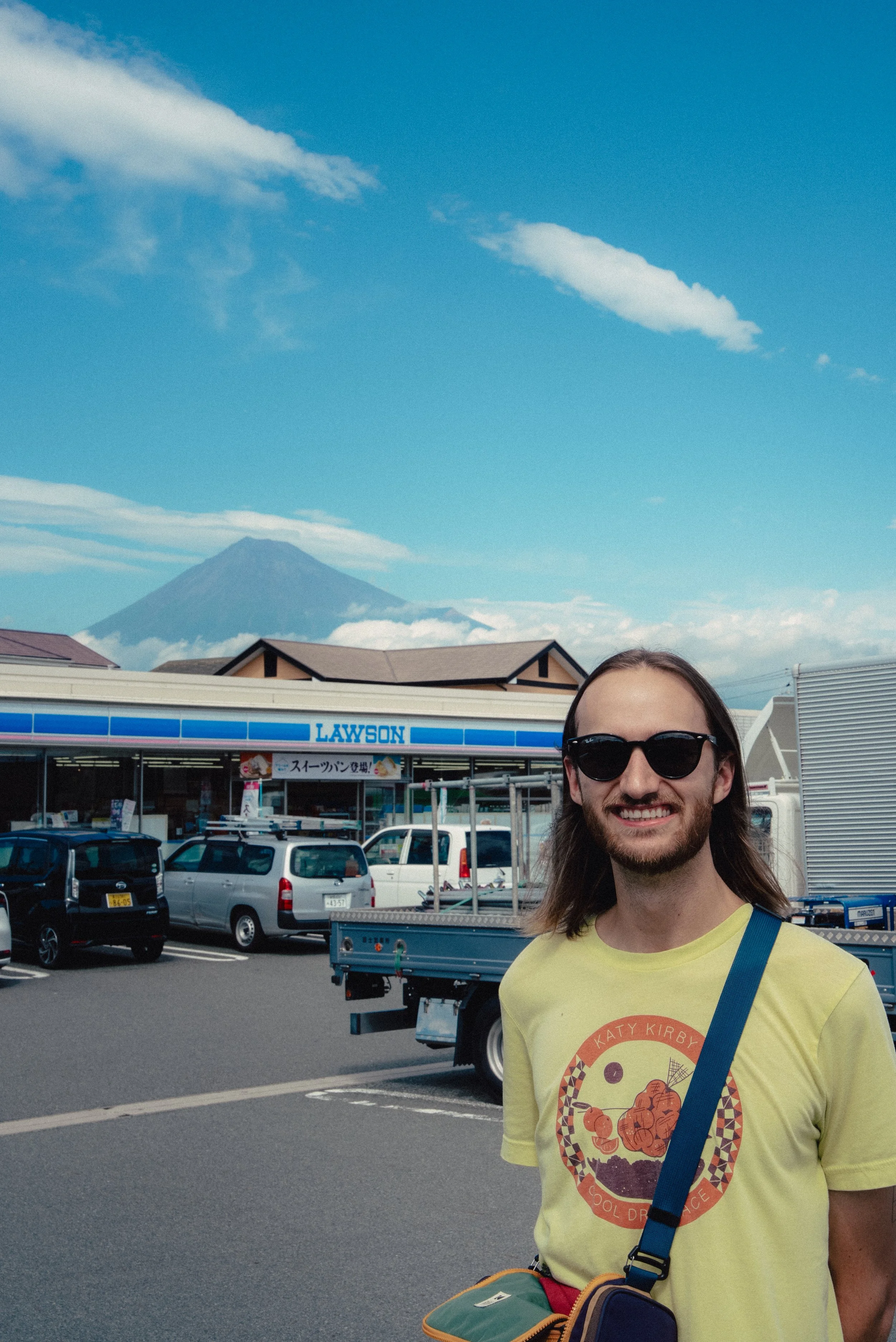 A man with long hair, beard, sunglasses, and a yellow T-shirt with a cartoon graphic, standing in a parking lot in front of a Lawson convenience store with Mount Fuji in the background.