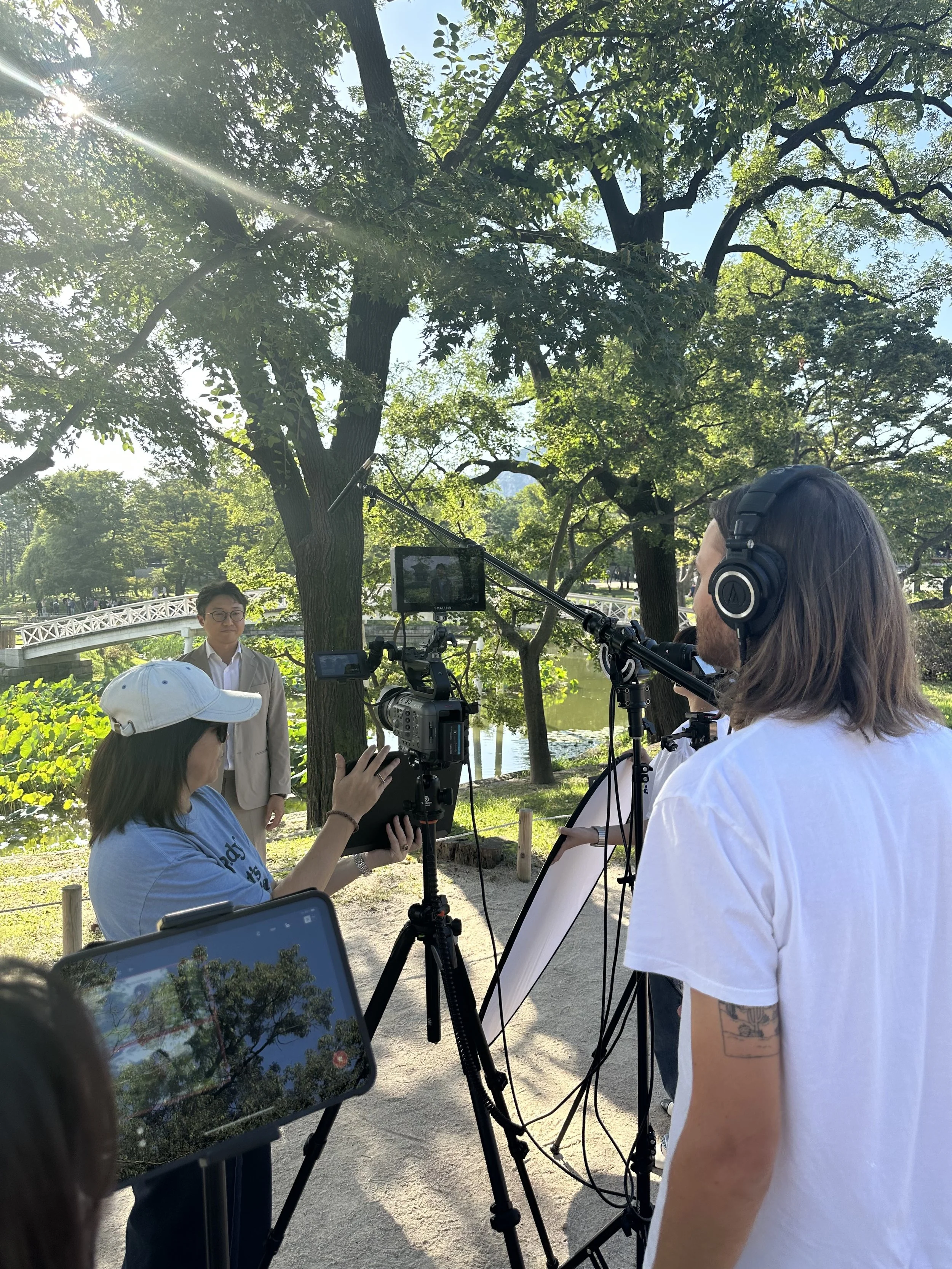 Filmmakers filming a person outdoors in a park near a pond with trees and a bridge in the background, sunny weather.