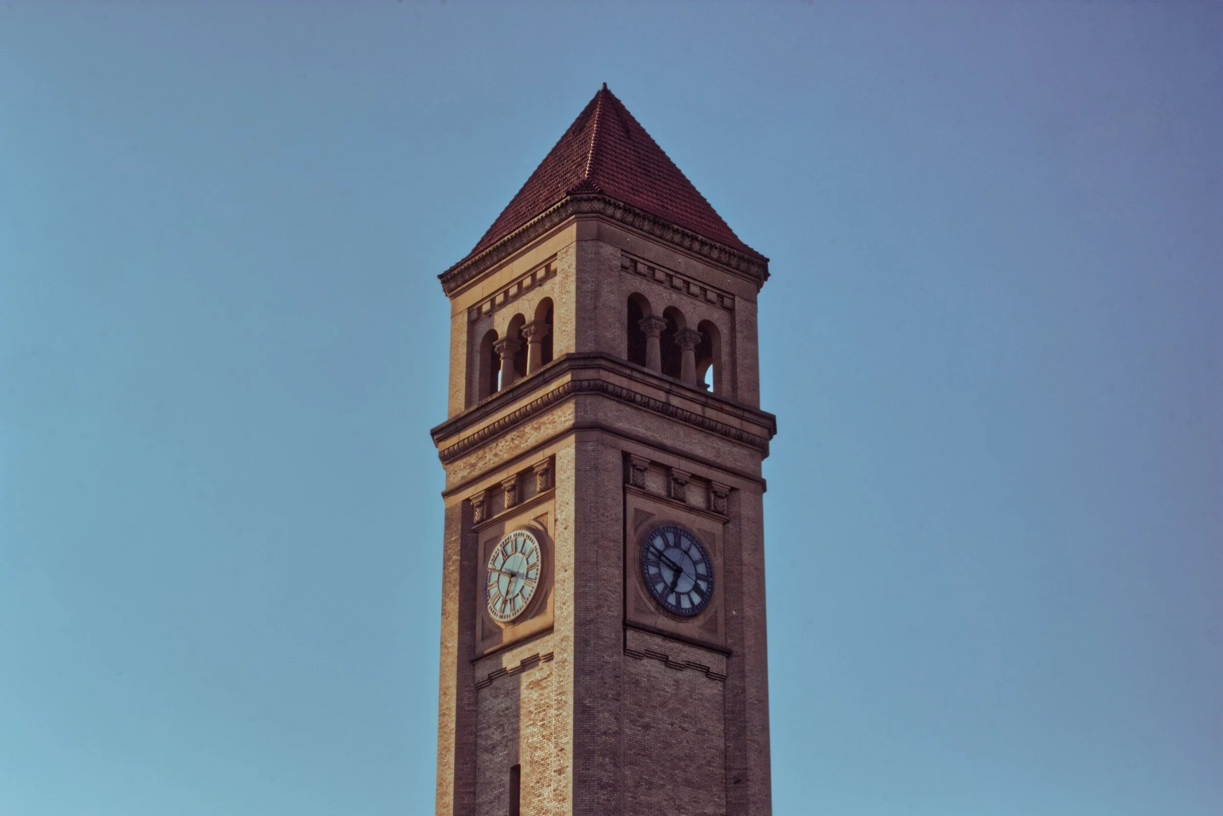 A tall clock tower with a red, pointed roof and a beige brick facade, set against a clear blue sky.