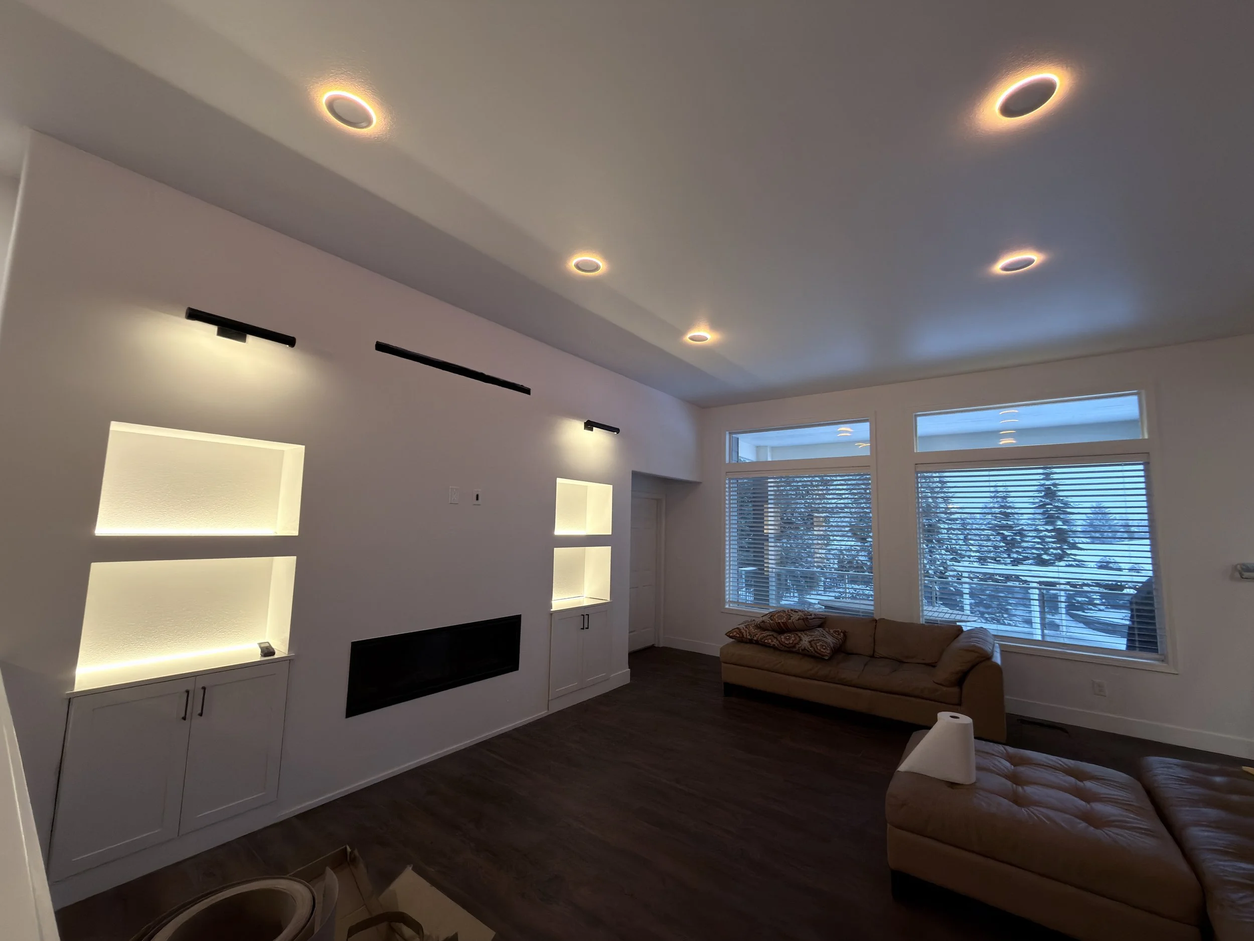 Living room with beige couch, large windows with blinds, modern ceiling lights, dark hardwood floors, and white built-in shelves with lighting.