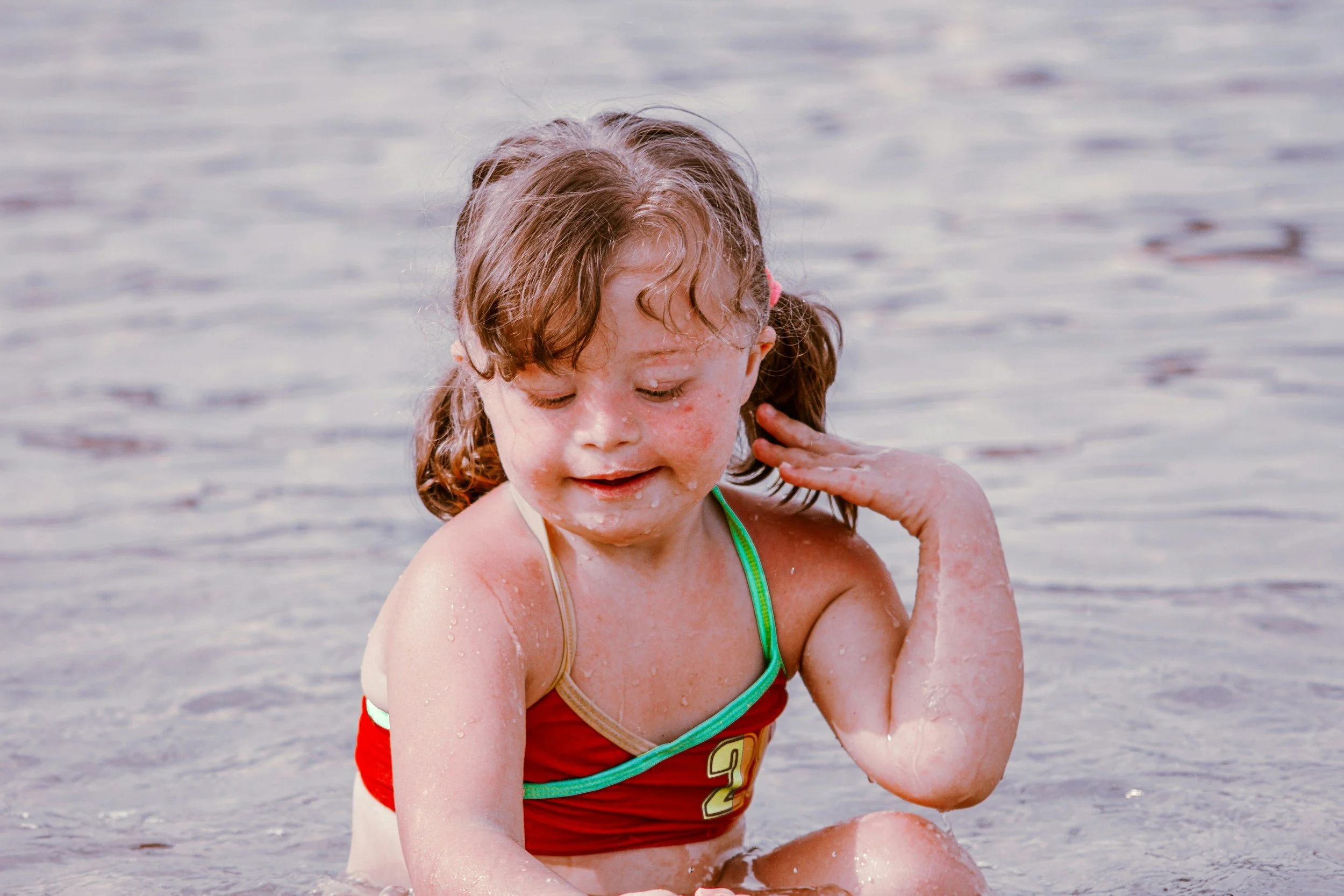 A young girl with curly brown hair is in shallow water at the beach, smiling and playing while wearing a red swimsuit with green trim.
