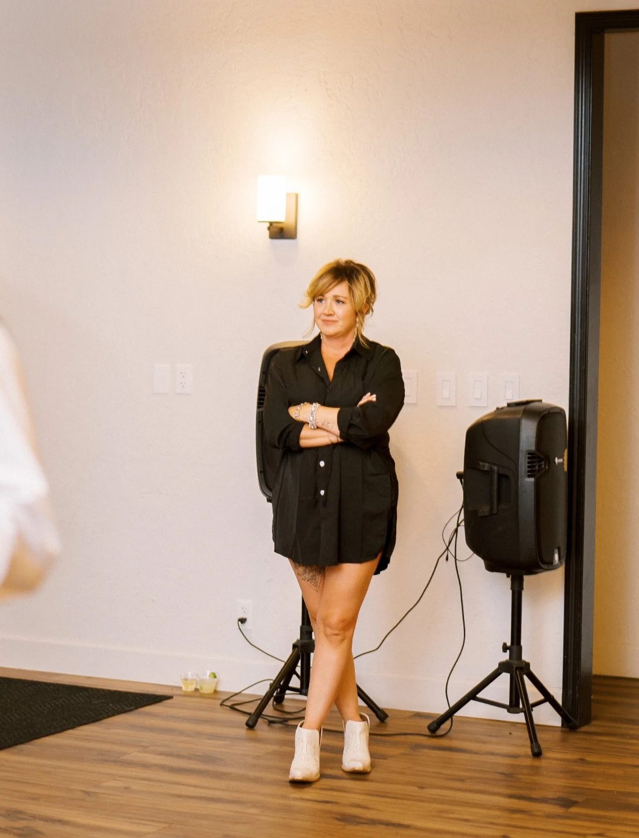 A woman with blonde hair standing in front of a plain wall, crossing arms, wearing a black shirt-dress and white cowboy boots, with speakers and audio equipment nearby.