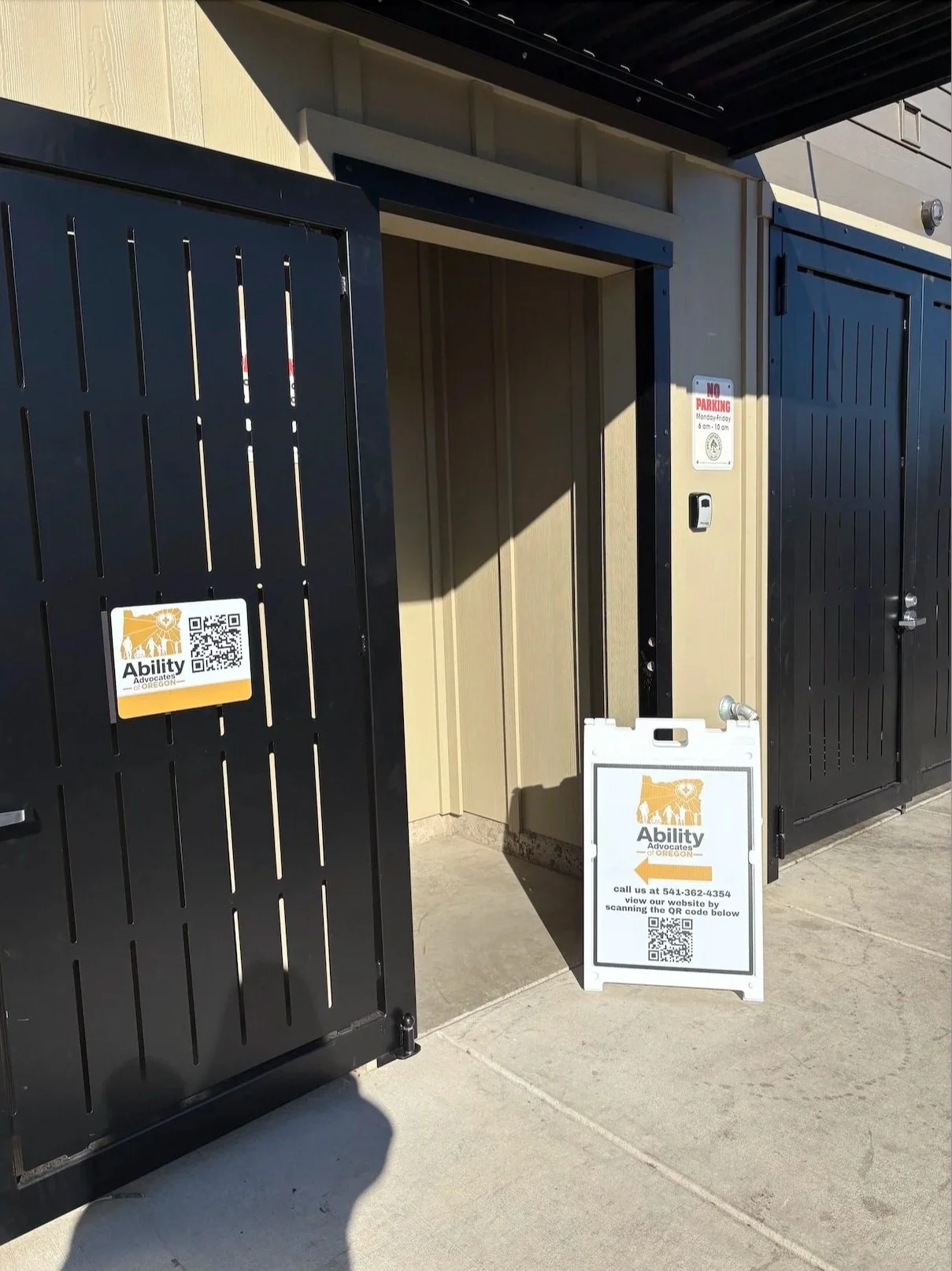 Outdoor area with two black metal gates, an open door, and an A-frame sign for Ability Advocates of Oregon, including a QR code and contact info.