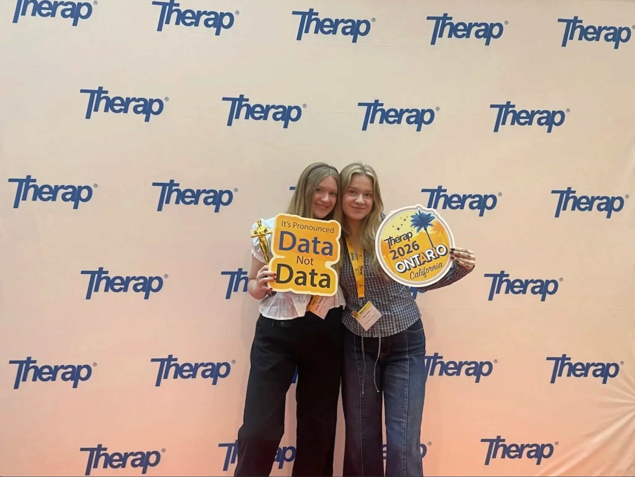 Two women standing together in front of a backdrop with the Therap logo, holding signs that say "It's Pronounced Data Not Data" and "Therap 2026 Ontario California."