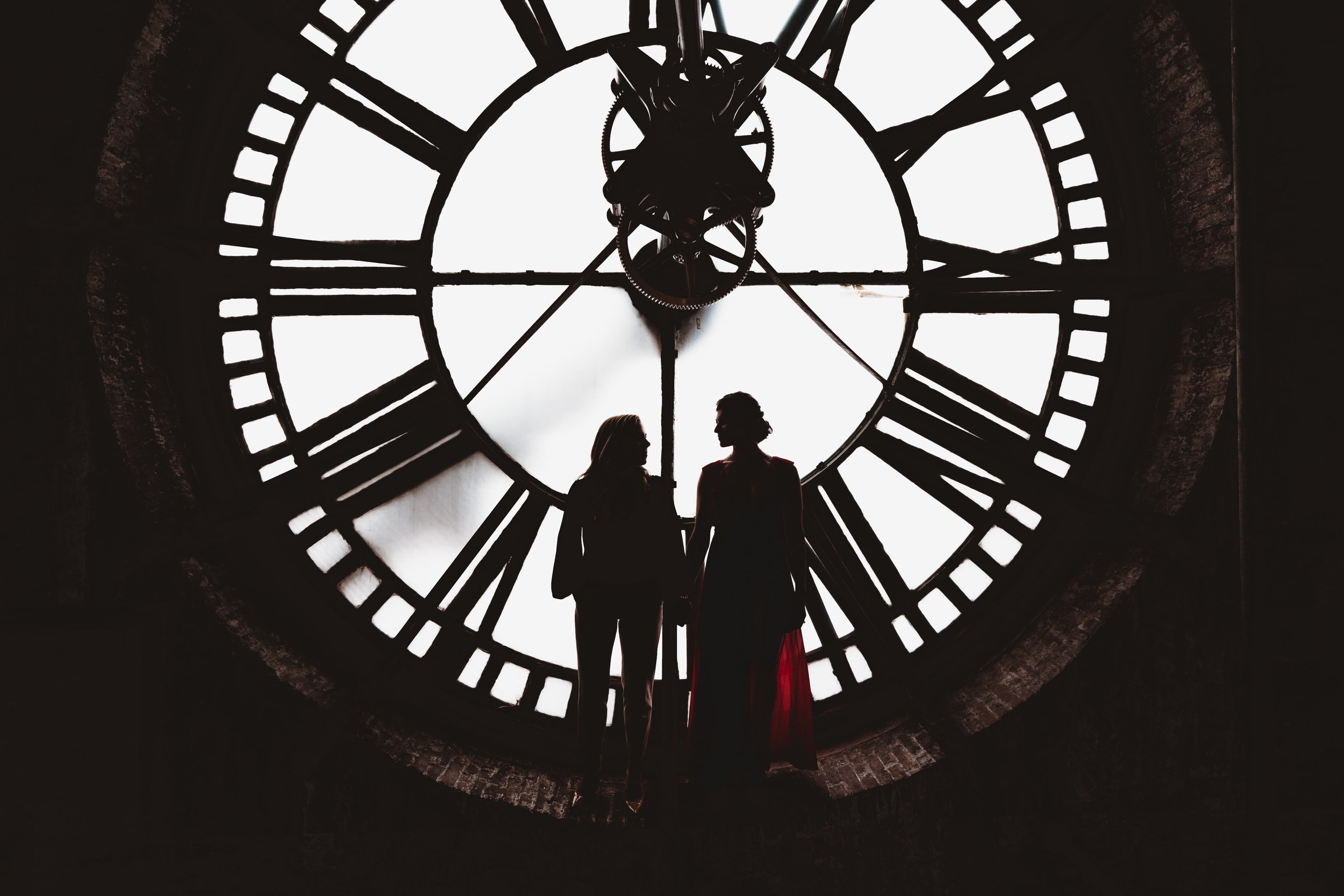Silhouettes of two people in front of a large clock face with Roman numerals, lit from behind.