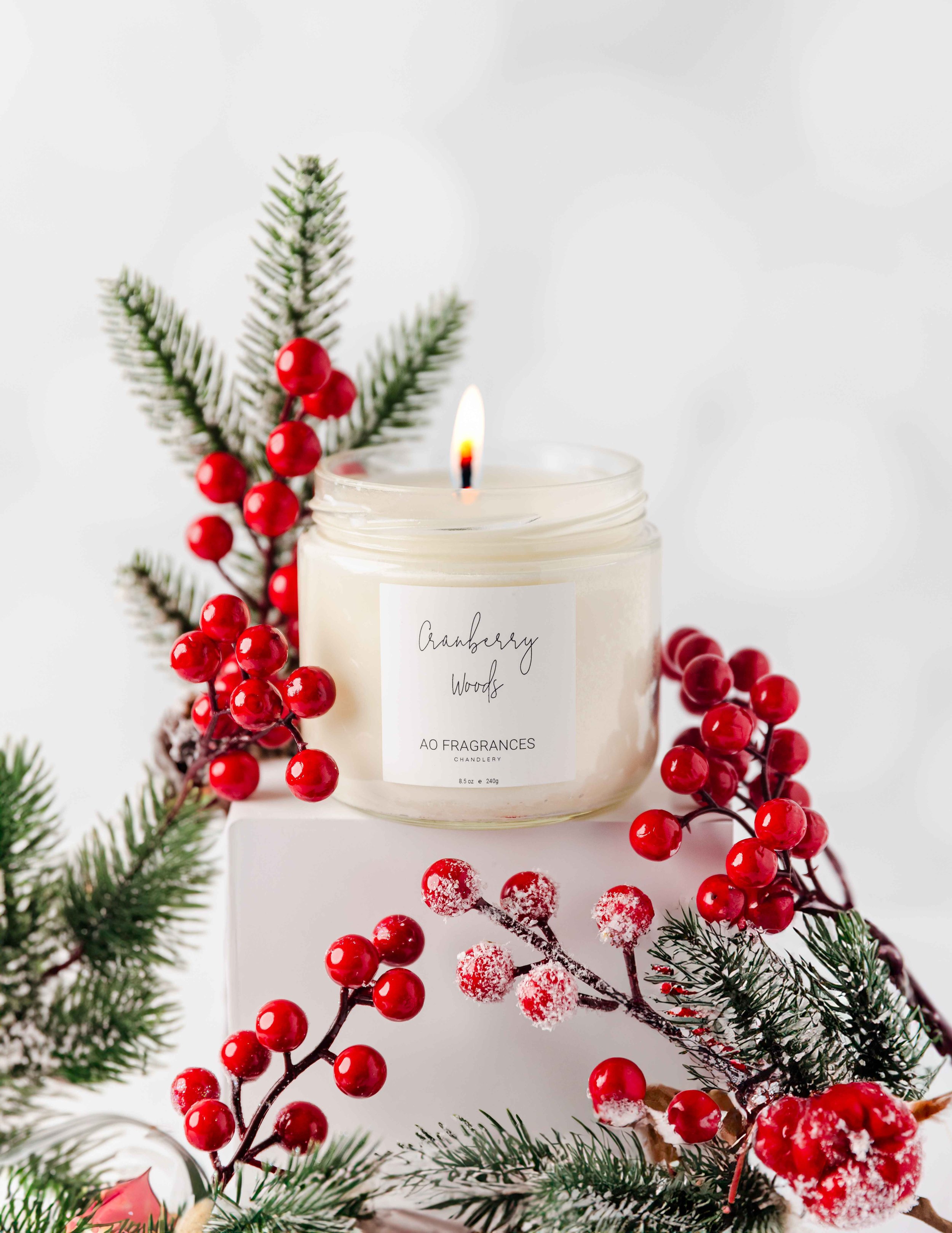 Lit candle in a glass jar labeled "Cranberry Woods" with evergreen branches and red berries.