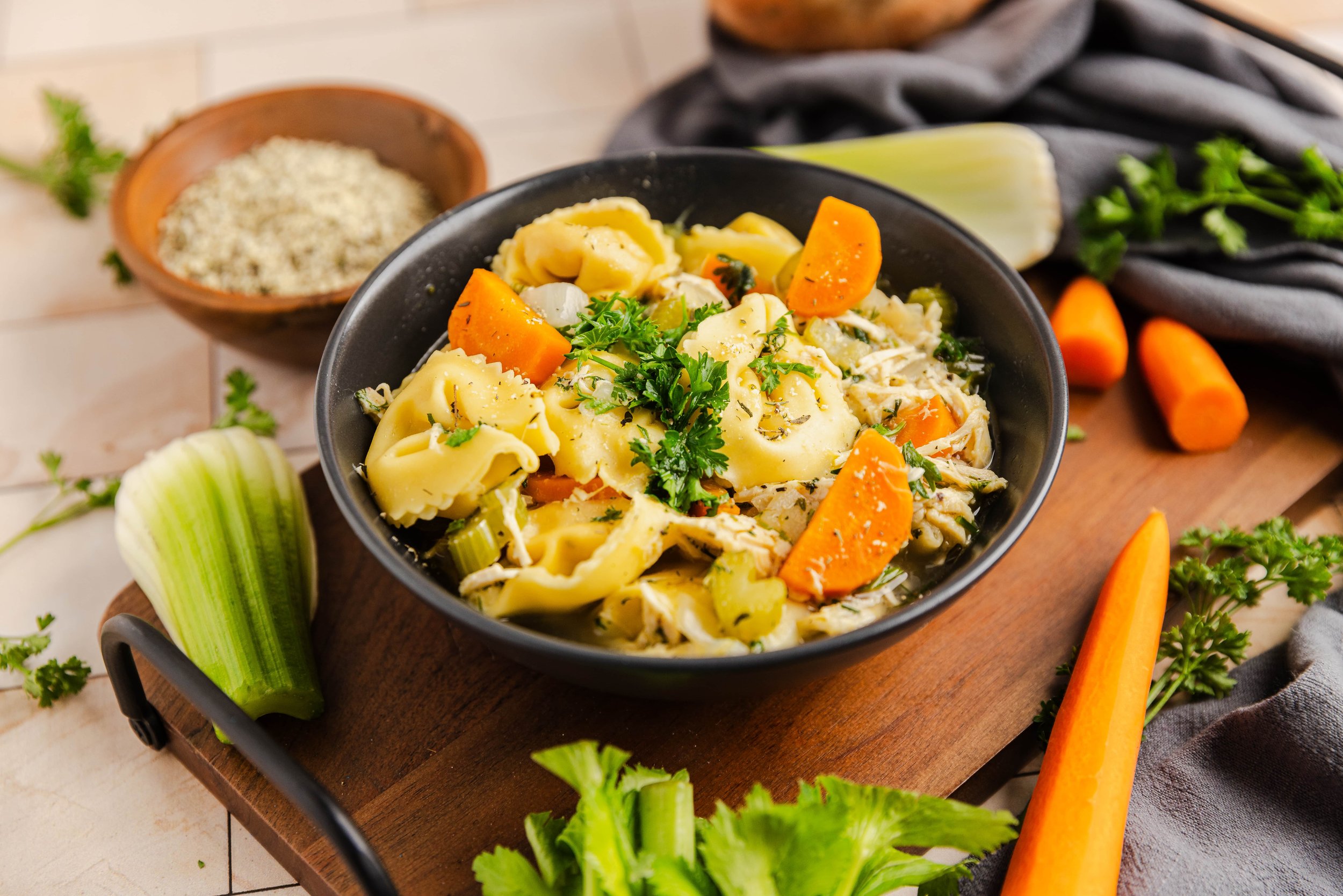 Bowl of tortellini soup with carrots, celery, parsley, and shredded chicken on a wooden board, surrounded by raw vegetables and herbs.