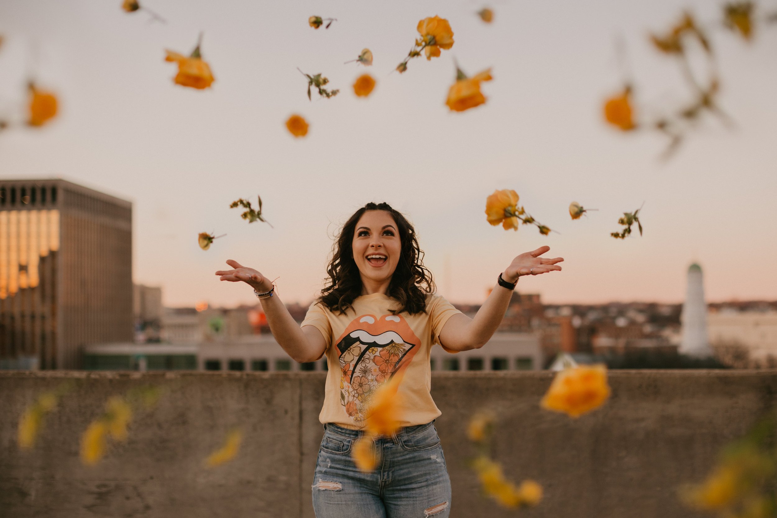 Woman in a t-shirt with a lip graphic, joyfully throwing yellow flowers in the air, standing in an urban setting during sunset.
