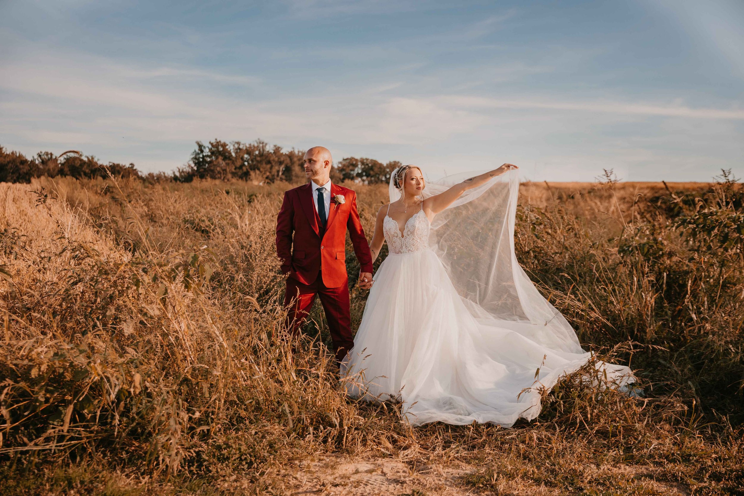 Bride and groom standing in a field, bride holding veil, groom in red suit, outdoor wedding photo.