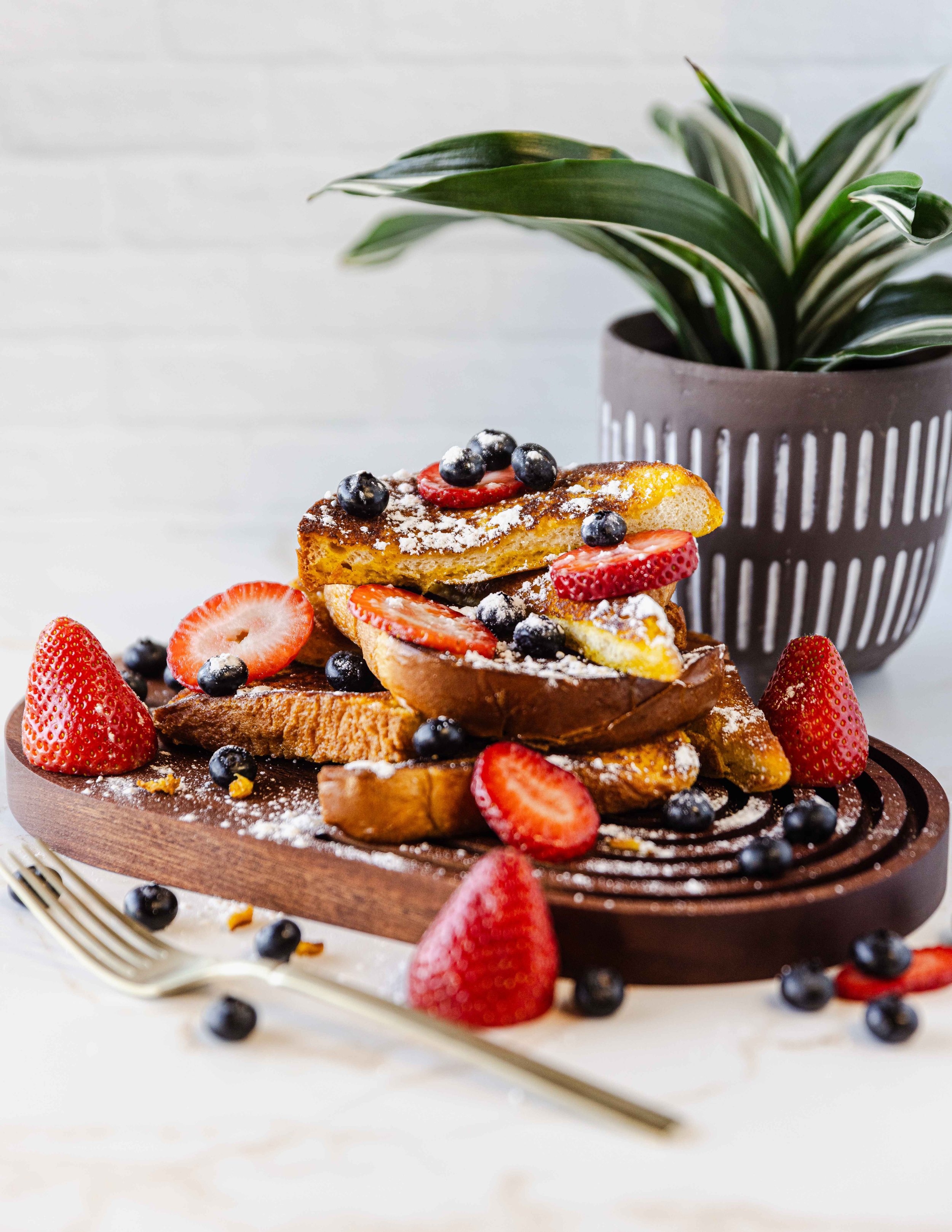 French toast with strawberries and blueberries on a wooden board, garnished with powdered sugar, beside a fork and a potted plant.