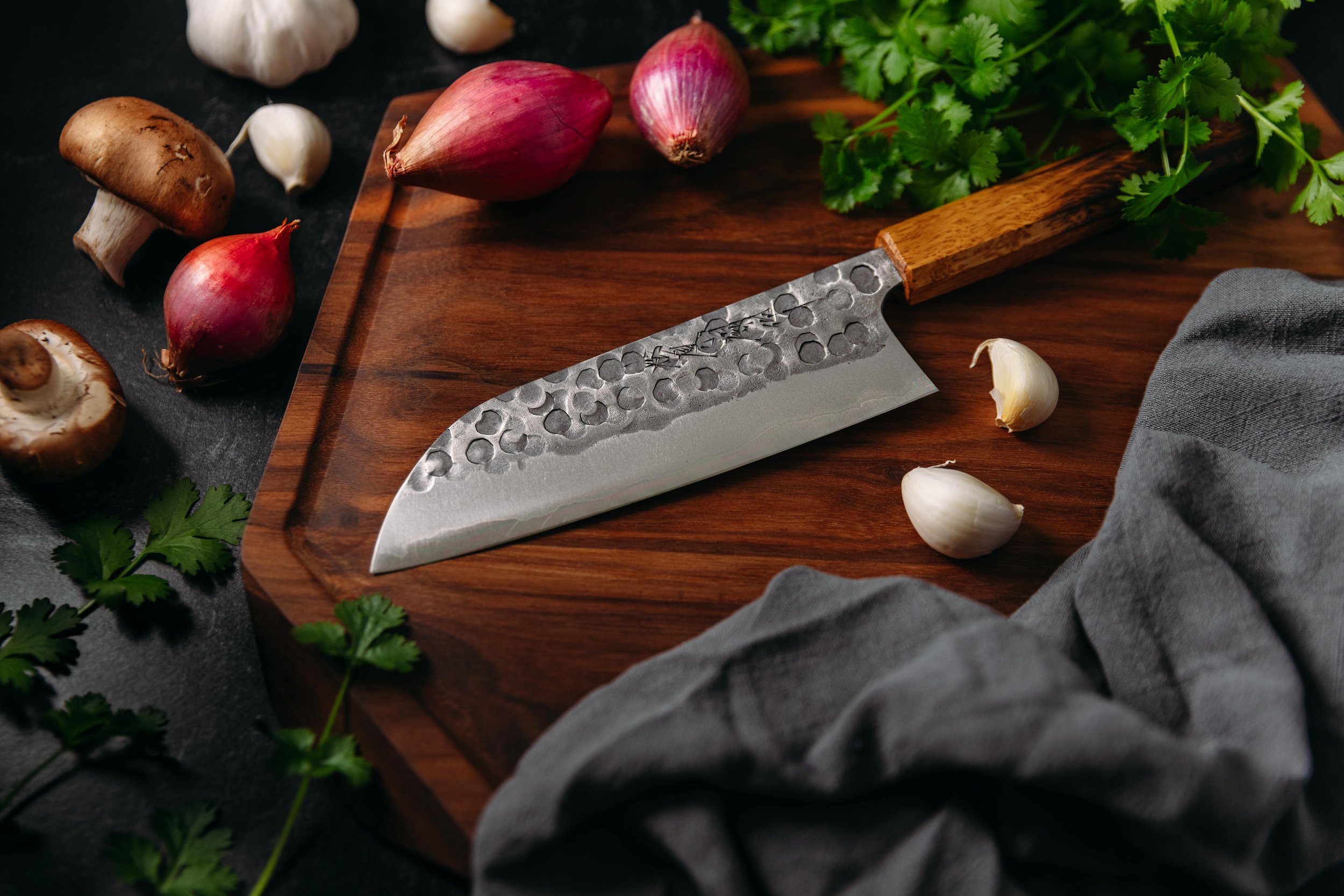 Japanese knife on wooden cutting board with mushrooms, red onions, garlic cloves, parsley, and gray cloth.