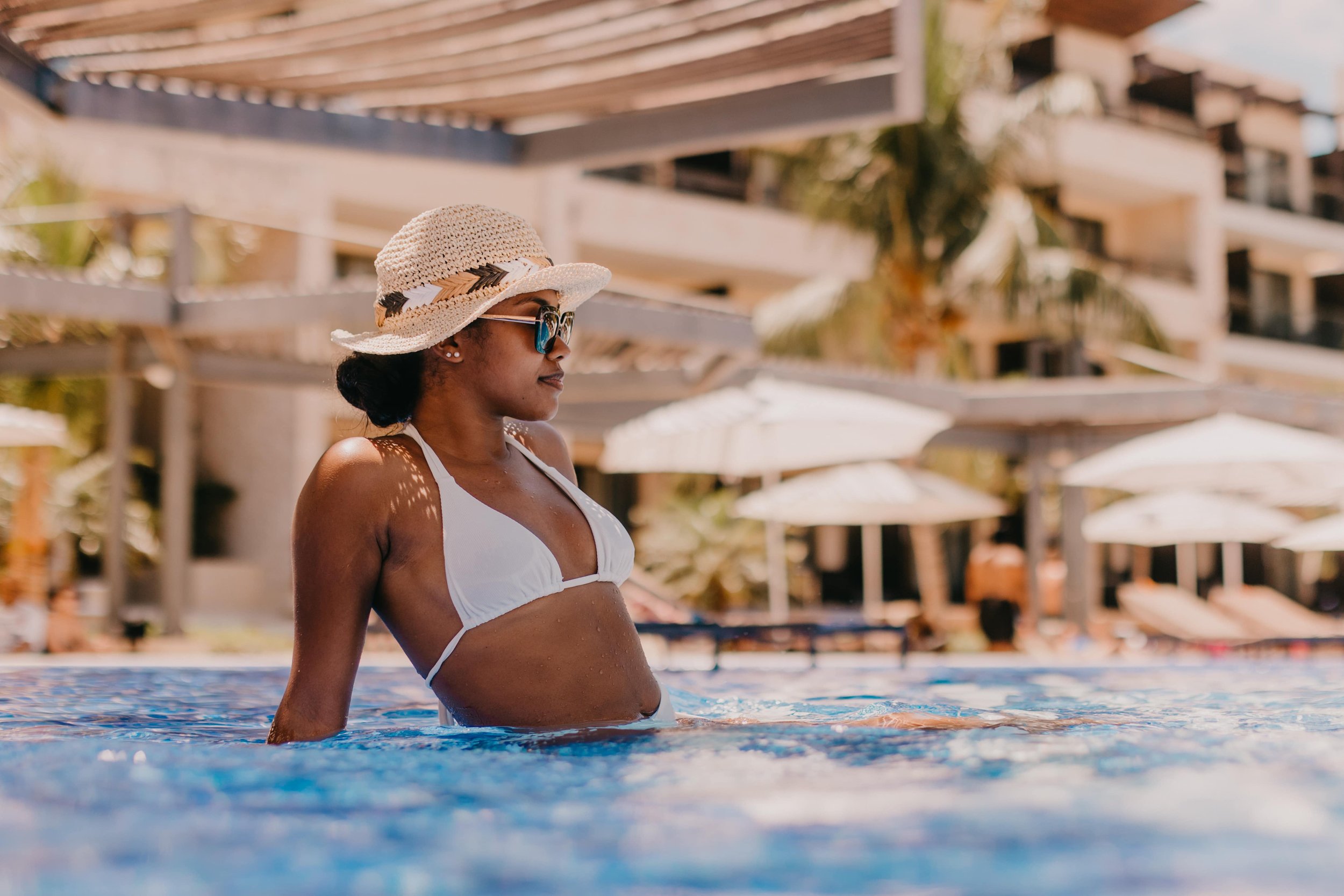 Woman in a straw hat and white bikini leaning on edge of a swimming pool, with resort buildings and umbrellas in the background.