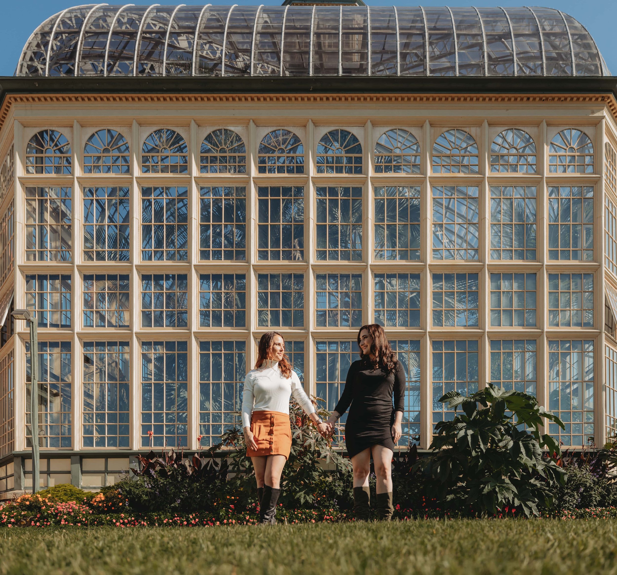 Two people walking in front of a large glass conservatory with a grid of windows, surrounded by greenery and flowers on a sunny day.