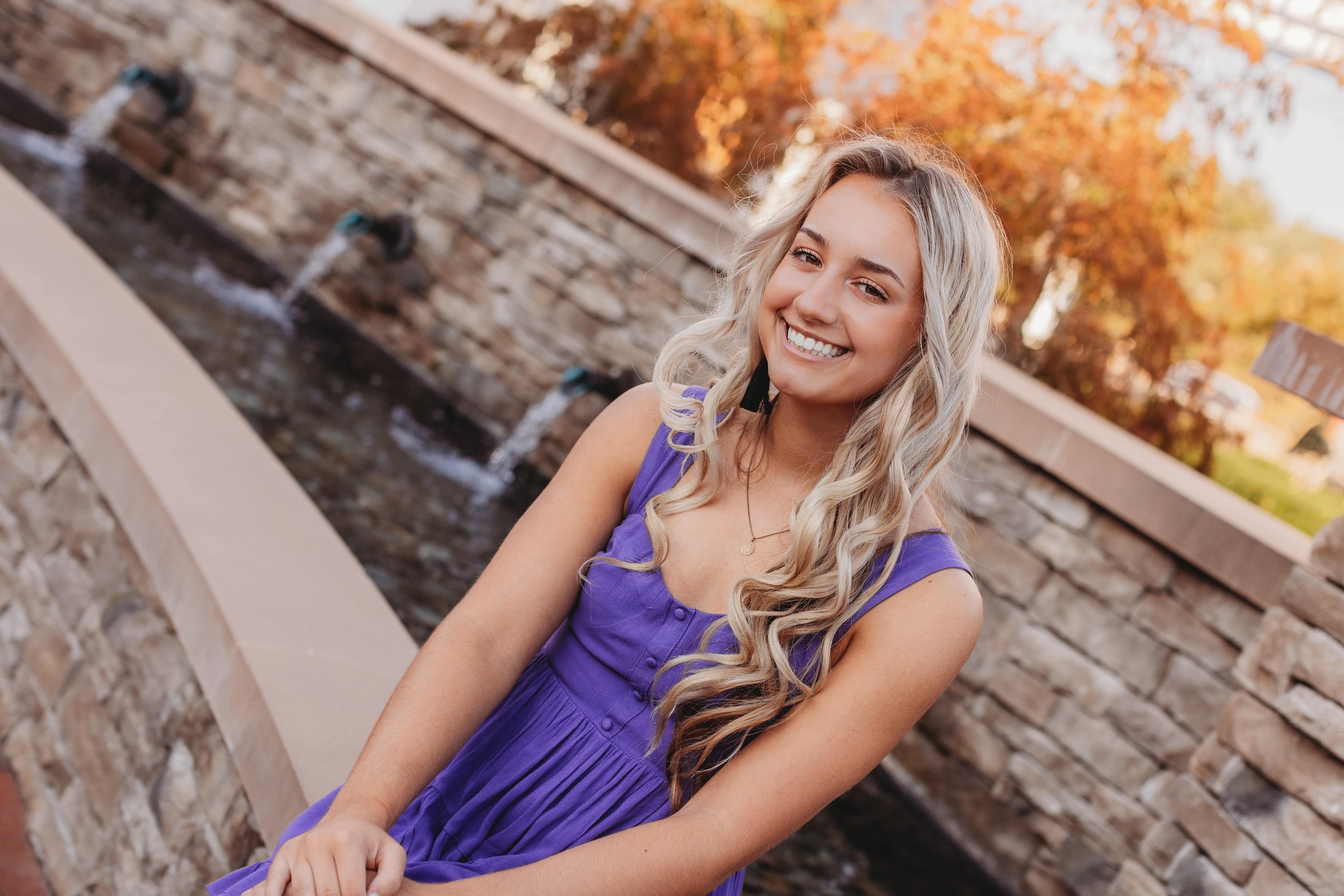 Smiling woman with long blonde hair wearing a purple dress sits by a stone fountain with water flowing, surrounded by autumn foliage.