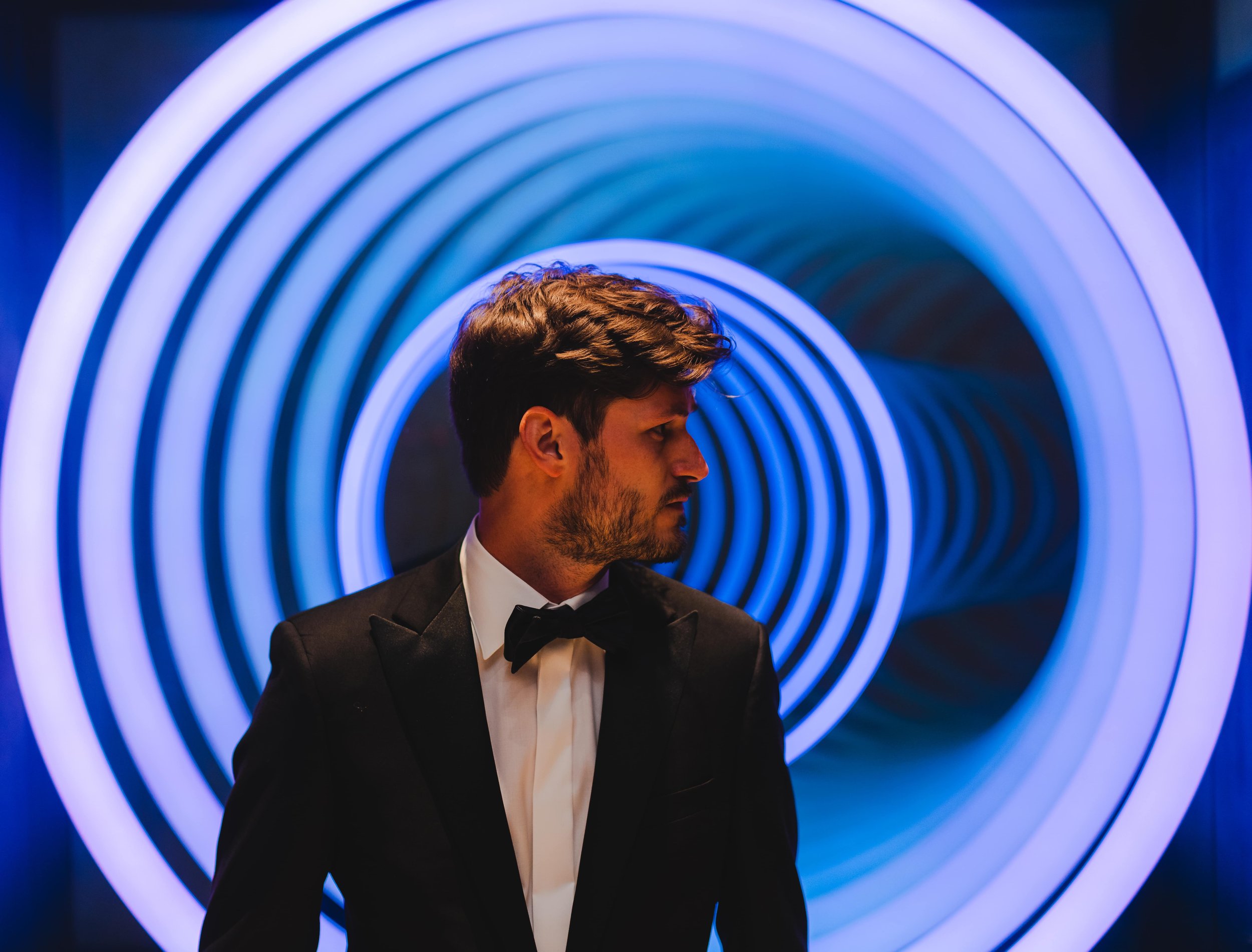 Man in tuxedo standing in front of concentric blue neon lights.