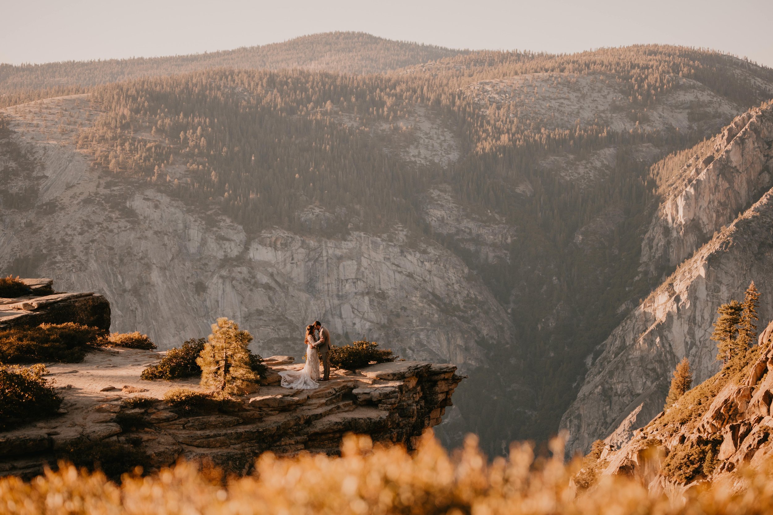 A couple in wedding attire stands on a rocky cliff with a mountainous landscape in the background, under warm, golden light.