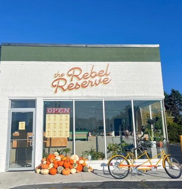 The Rebel Reserve storefront with a yellow tandem bicycle parked outside and a pile of pumpkins at the entrance, under a clear blue sky.