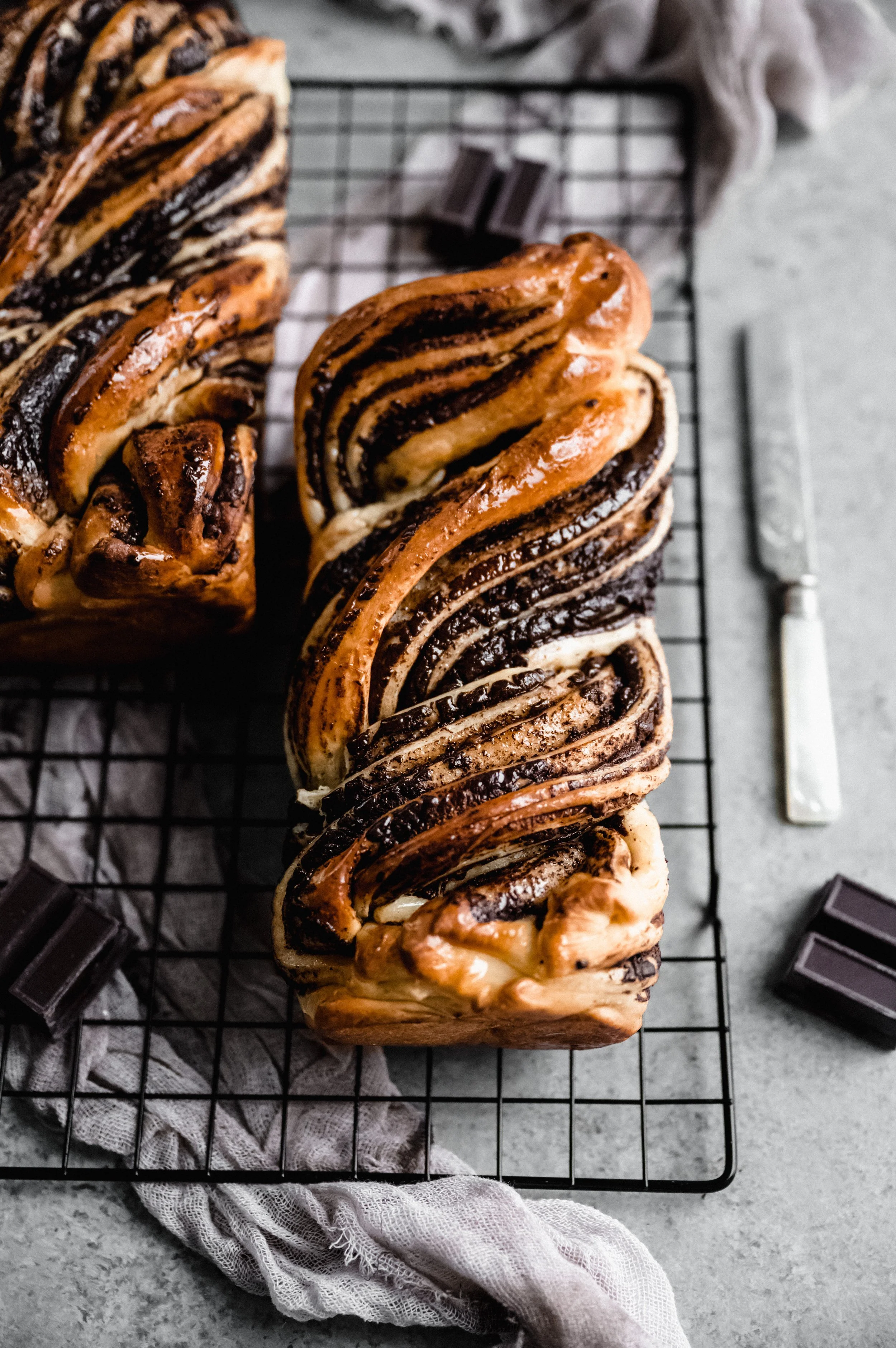 Chocolate babka bread on a cooling rack with pieces of chocolate and a knife nearby.