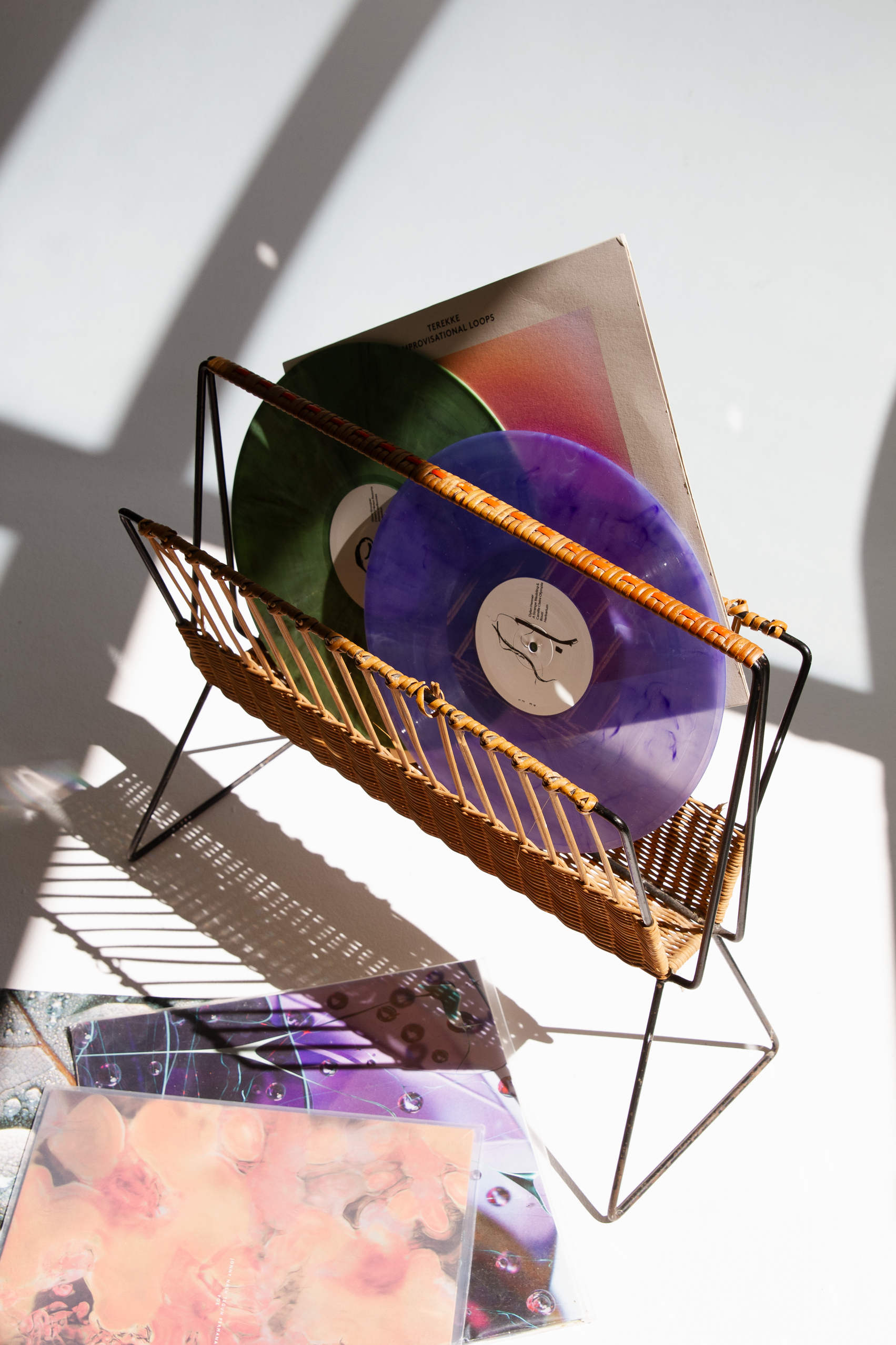 A metal and wicker magazine holder with multicolored vinyl records inside, placed on a white surface with shadows cast by sunlight, and some colorful vinyl record covers in the foreground.