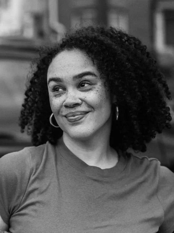 A woman with curly hair and hoop earrings, smiling and looking to her left, wearing a casual t-shirt.