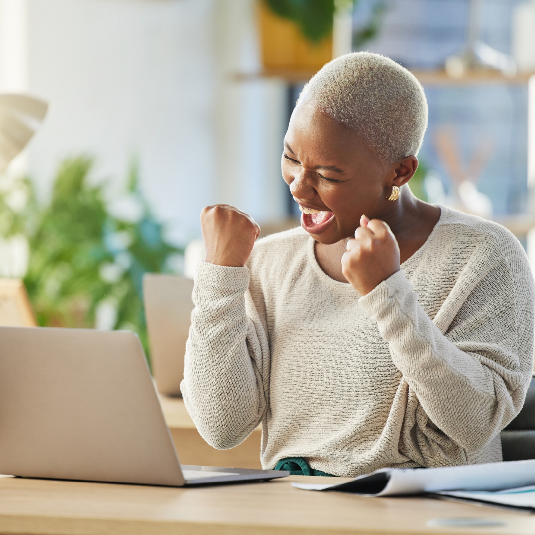 A woman with short blonde hair celebrating with clenched fists and a joyful expression while sitting at a desk with a laptop and documents.