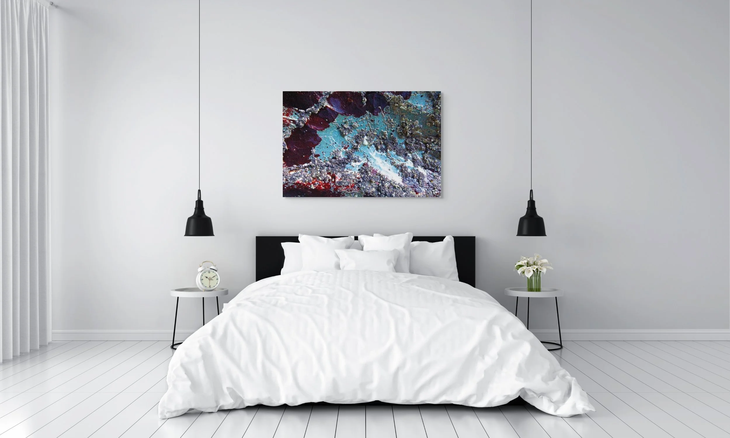 Minimalist bedroom with white walls and floor, black headboard, white bedding, two small white tables on each side, one with flowers, the other with a clock, two black hanging lamps, and abstract art on the wall.