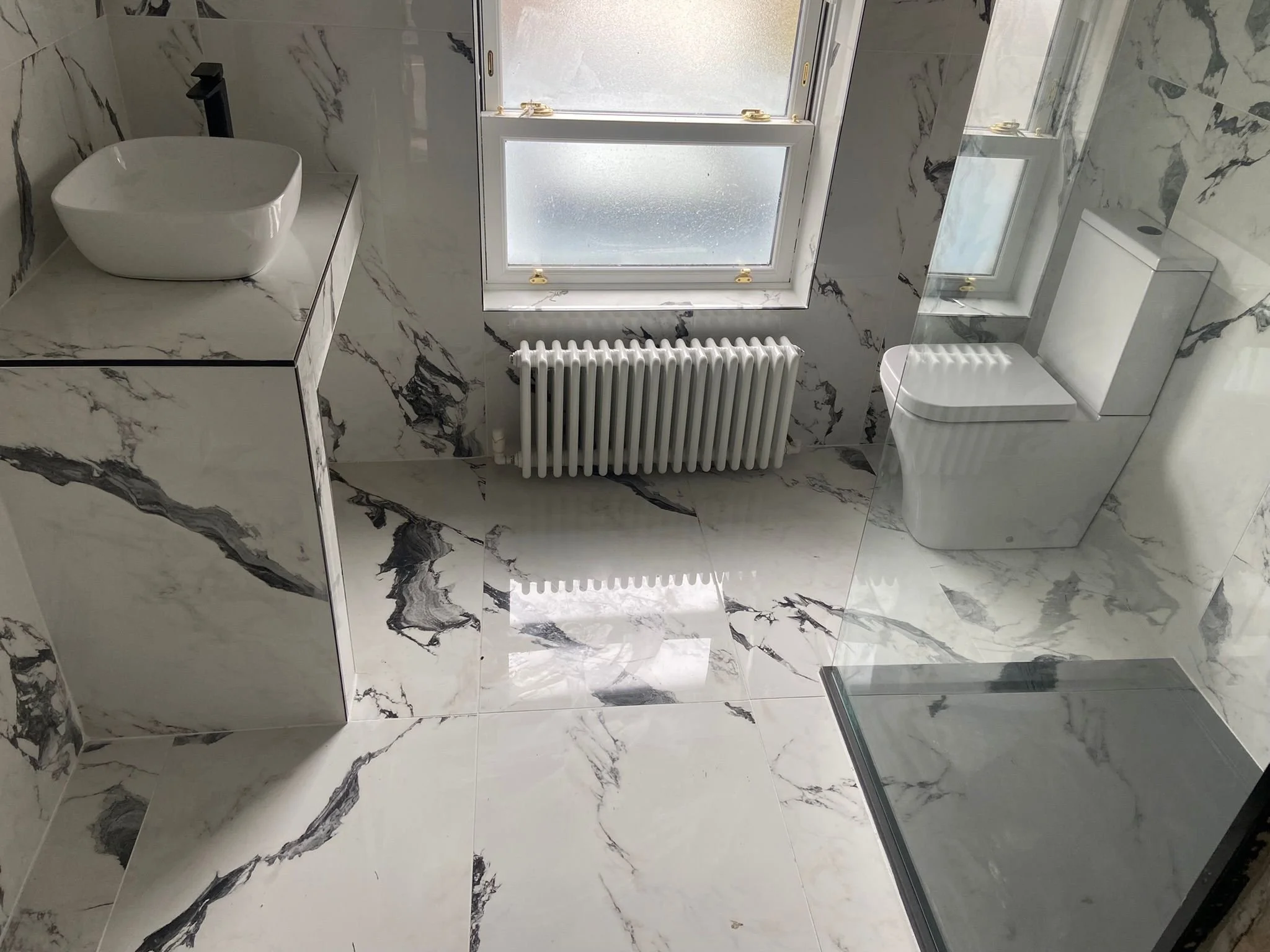 Modern bathroom with marble walls and floor, a frosted window, a white vessel sink on a marble countertop, a black faucet, radiators beneath the window, and a glass shower enclosure.