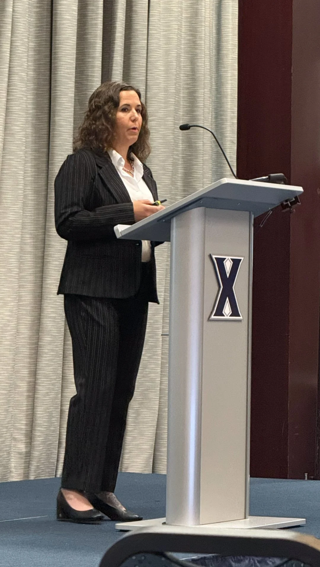 A woman in a black pinstripe suit giving a presentation at a podium with a Yale logo, standing in front of curtains.