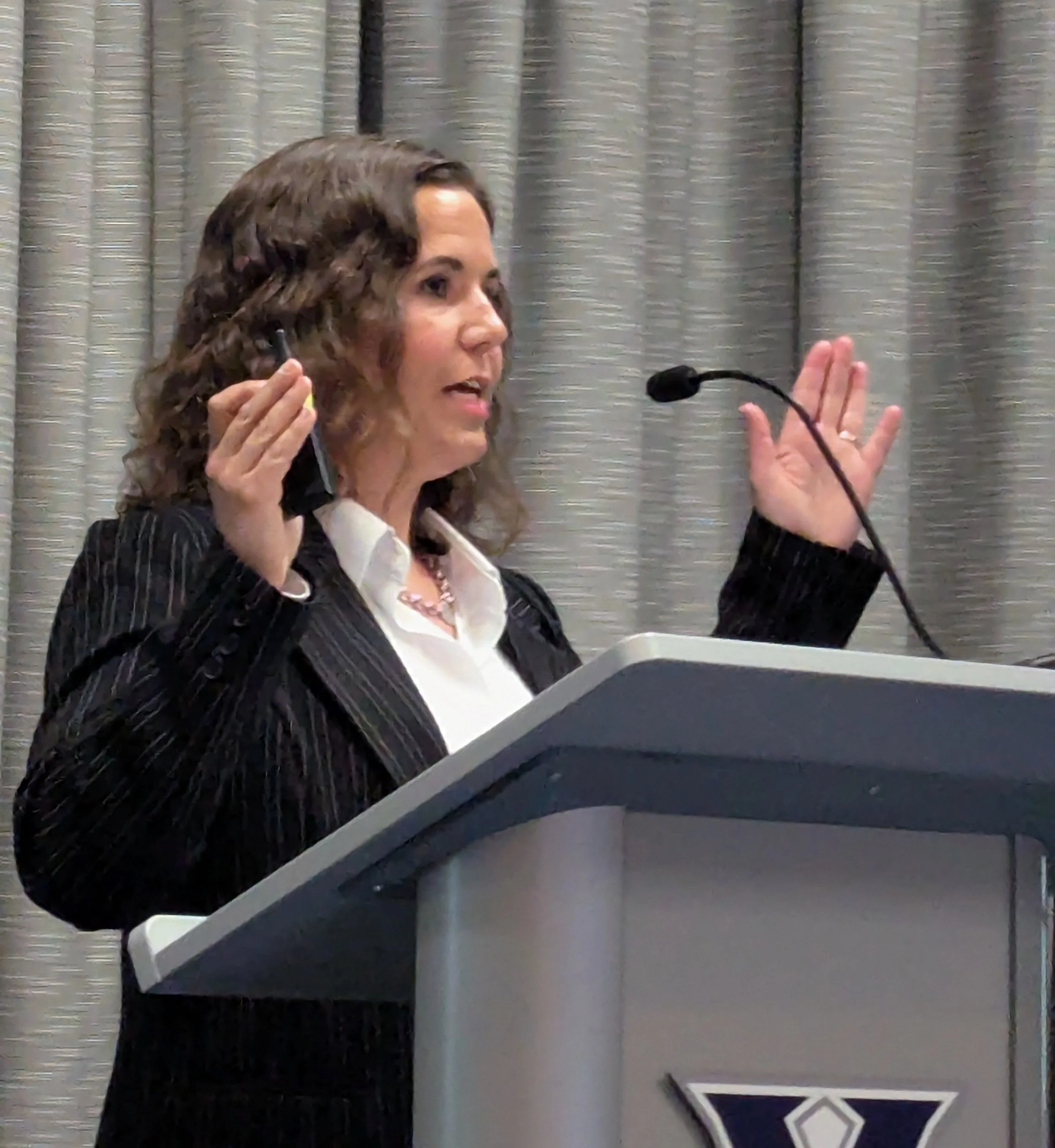 A woman with curly brown hair wearing a black blazer and white blouse speaking at a podium with a microphone, gesturing with her right hand, in front of a gray curtain.