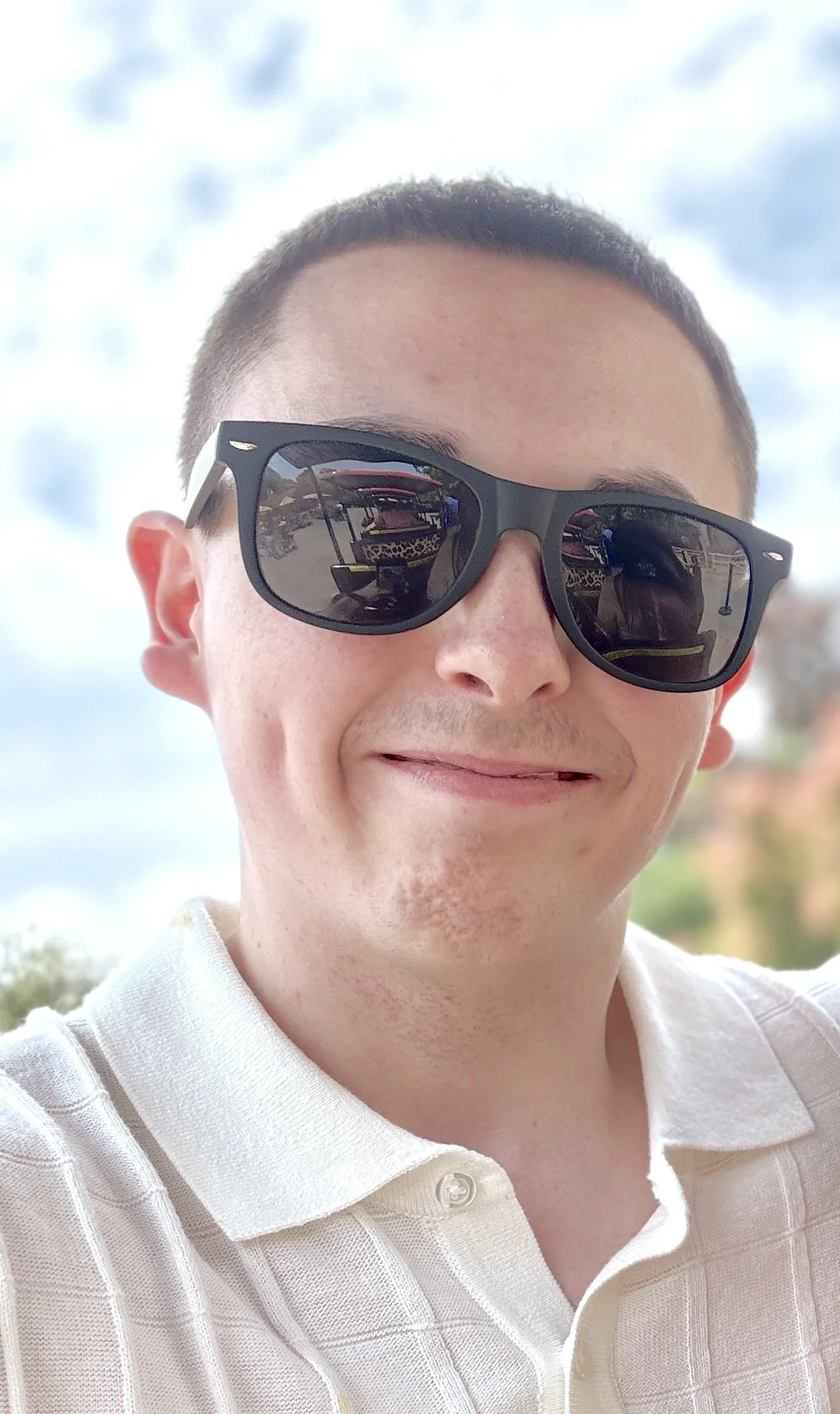 Close-up of a smiling person wearing sunglasses and a light-colored collared shirt, outdoors on a cloudy day.