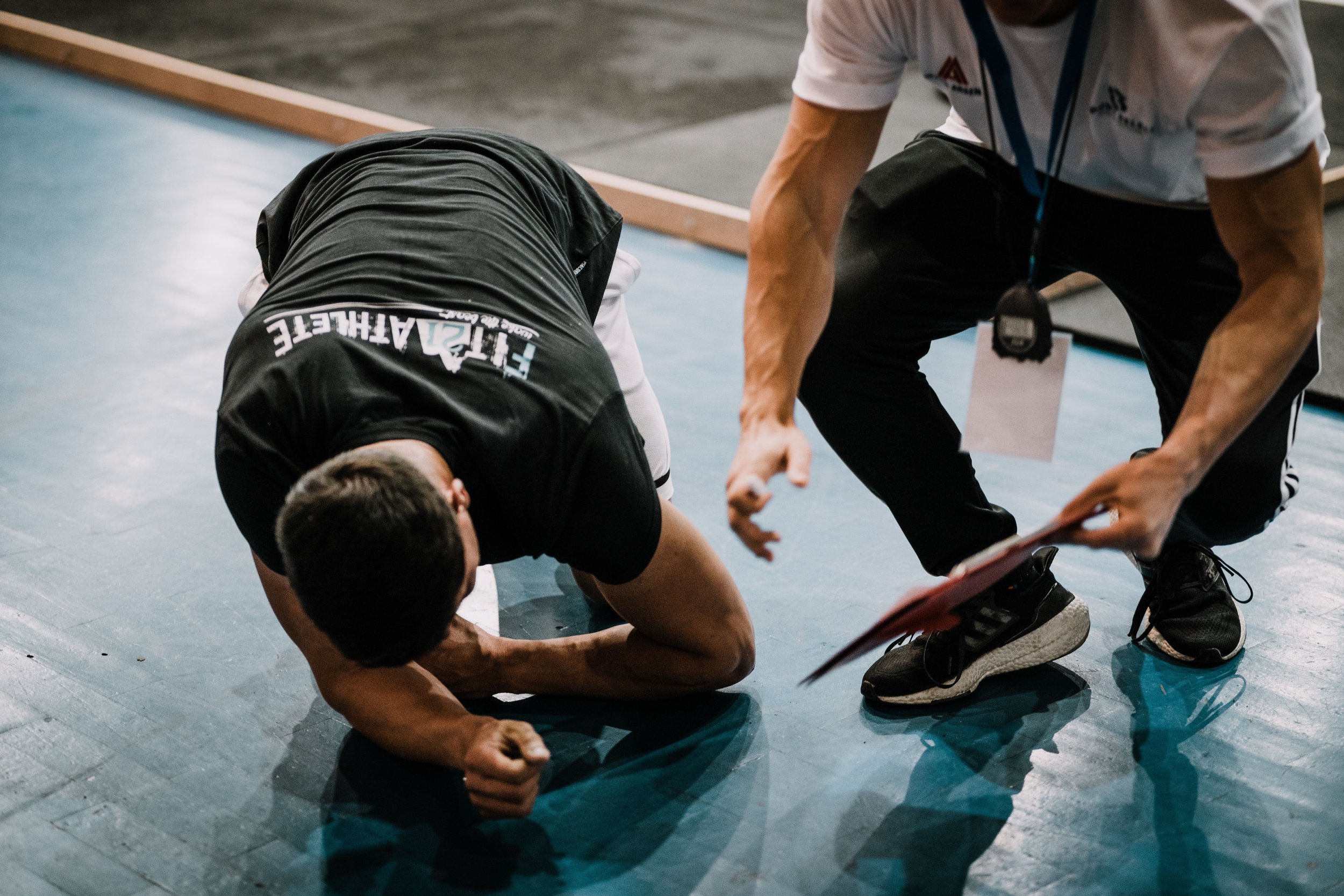 A man in sports attire is on the floor, seeming to be injured or exhausted, while another man wearing a white shirt and black pants is kneeling beside him, possibly providing assistance or checking on him.